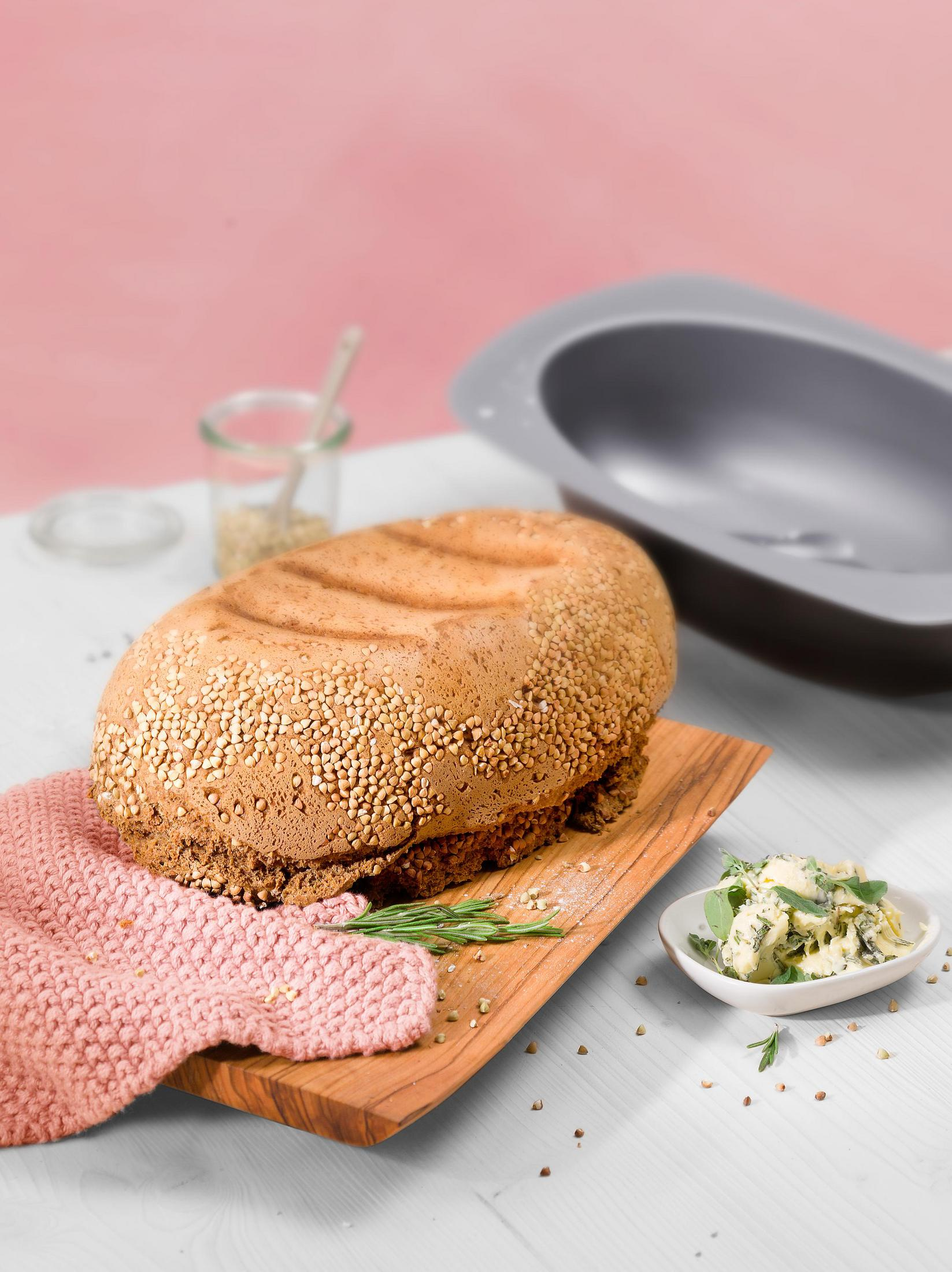 Brot mit Körnern auf einem Holzbrett, daneben Kräuterbutter und ein graues Objekt.