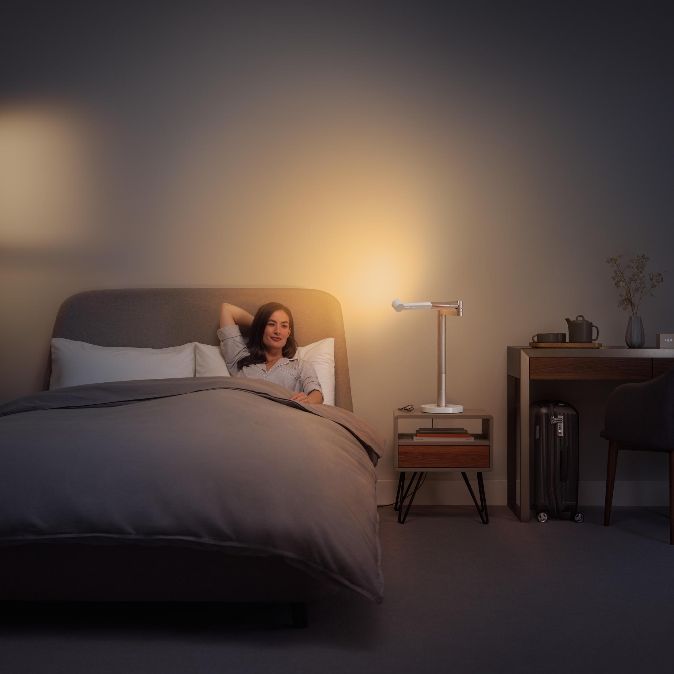 Frau im Bett liegend, Nachttisch mit Lampe, Schreibtisch mit Koffer und Stuhl.