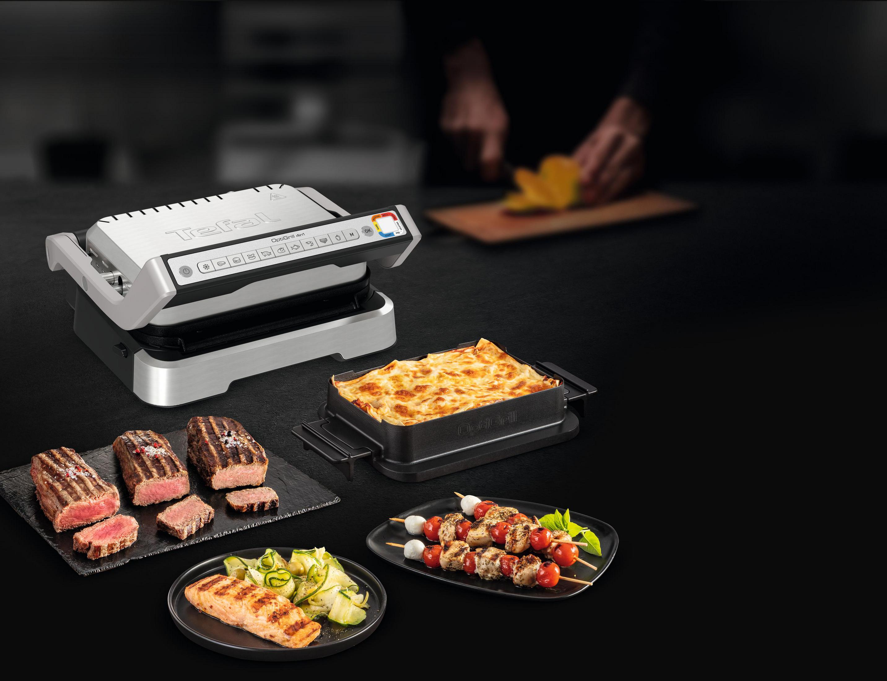 Ein silberner Kontaktgrill mit gekochten Speisen daneben: Steak, Lachs, Spieße und Lasagne.