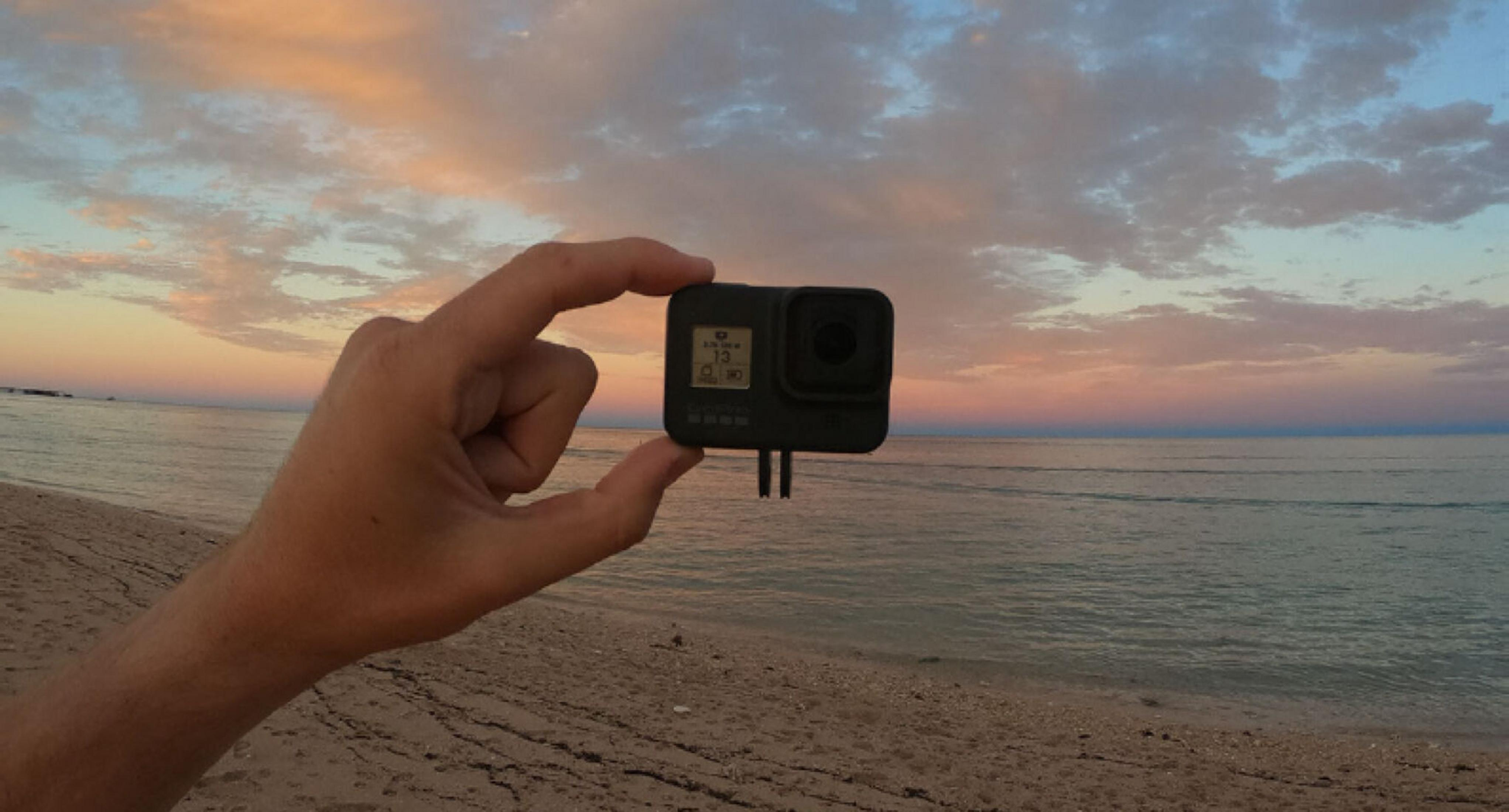 Hand hält GoPro vor Strand und Meer bei Sonnenuntergang.