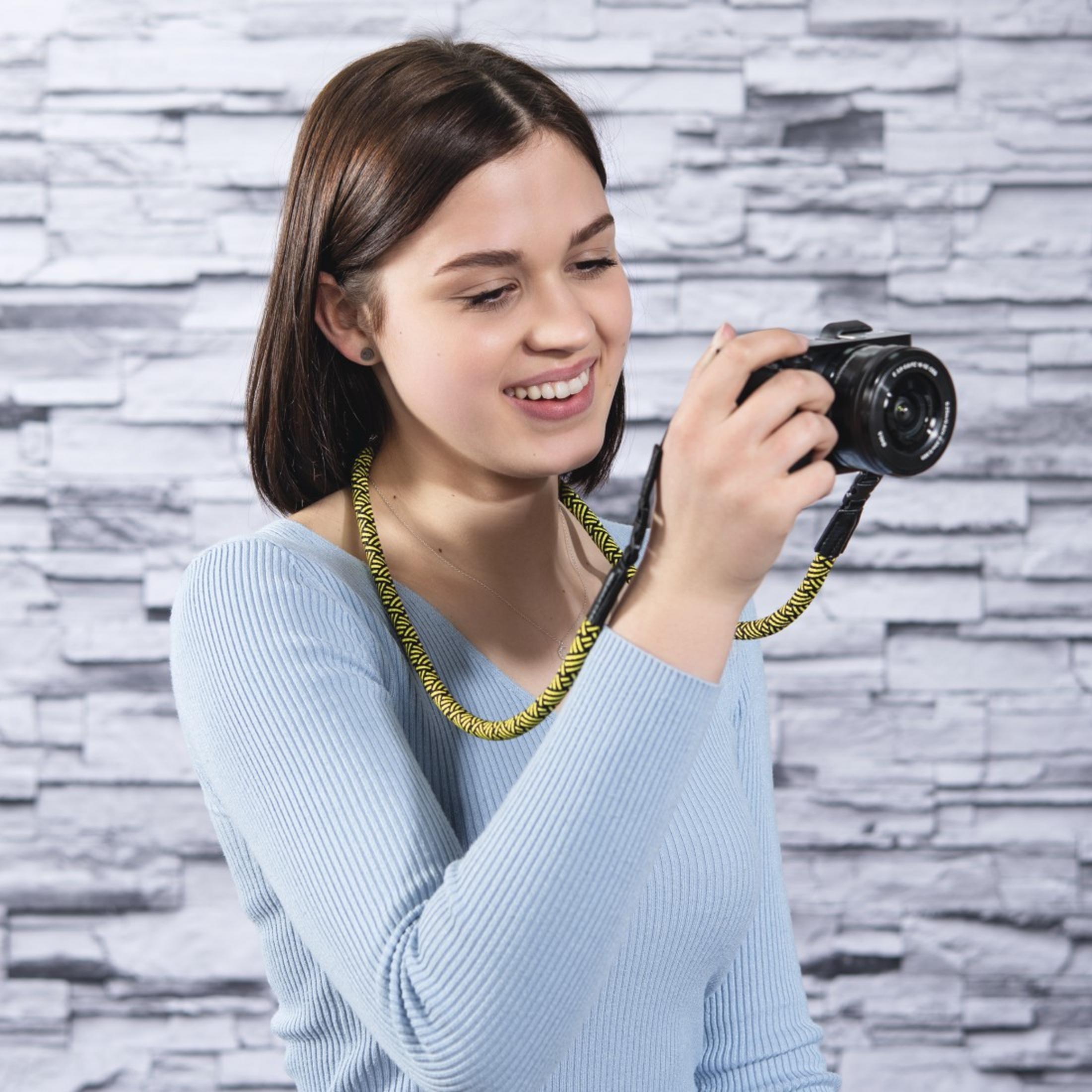 Młoda kobieta z brązowymi włosami trzyma aparat. Nosi niebieski top.