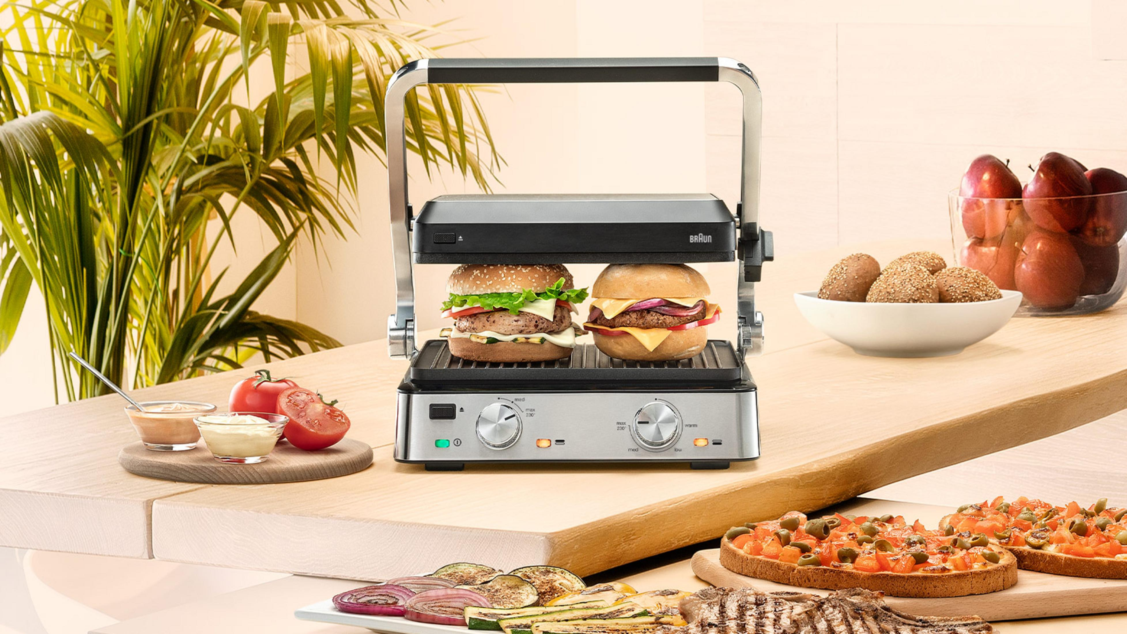 Grill kontaktowy Braun z dwoma burgerami, obok owoców, chleba i grillowanych warzyw.