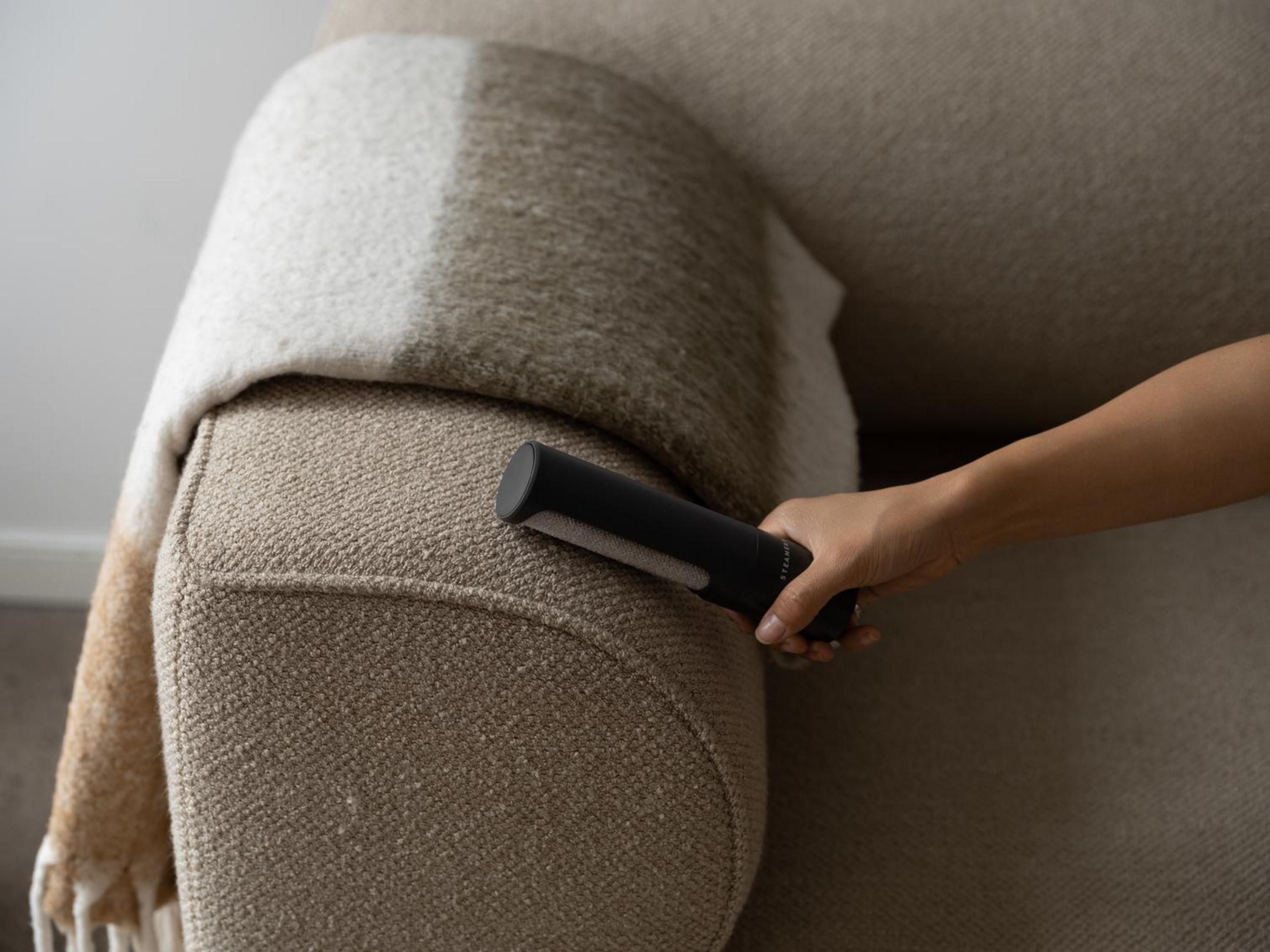 Eine Hand hält einen schwarzen Fusselentferner auf einem beigefarbenen Sofa mit einer Decke.