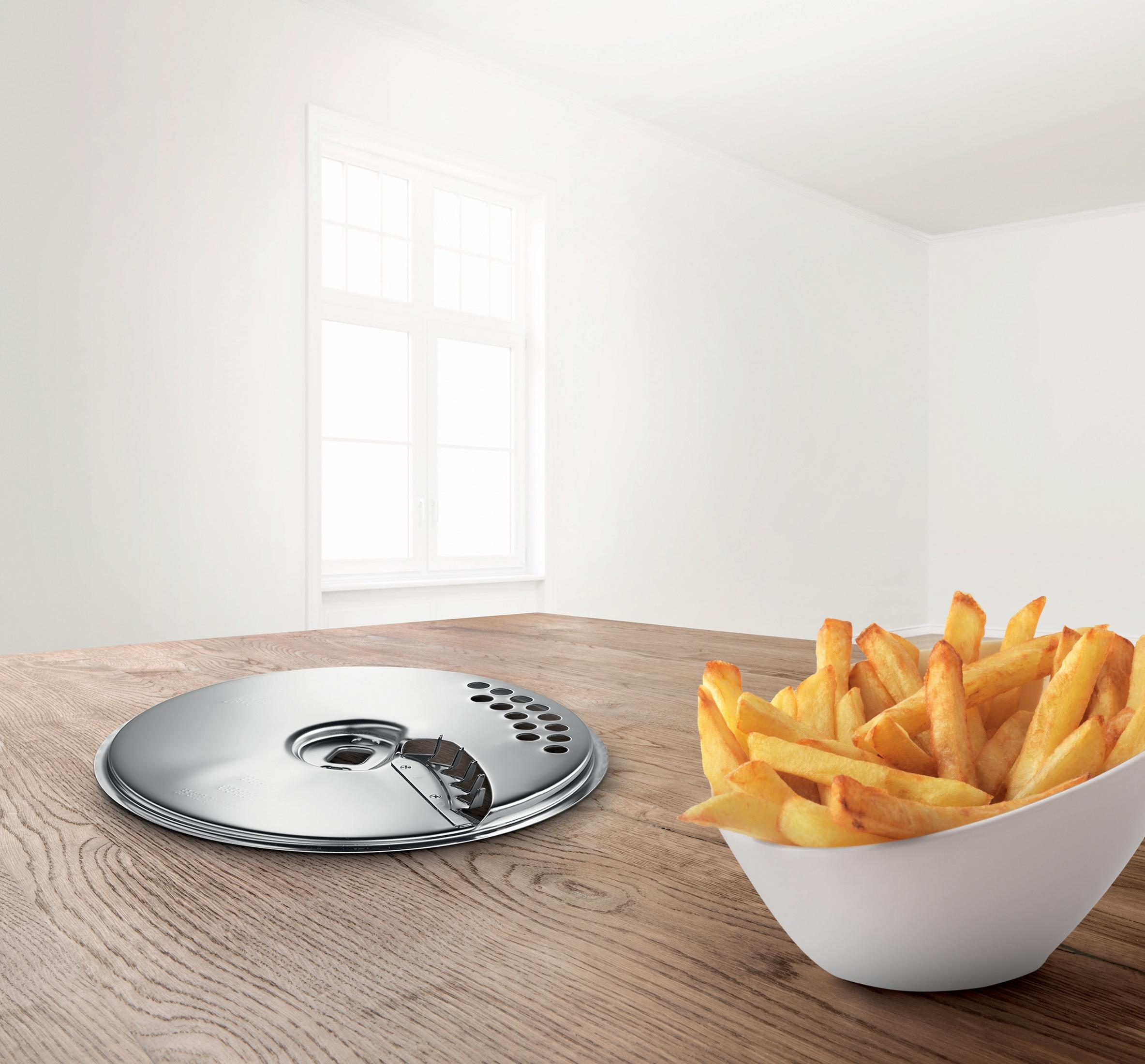 Ein metallischer Schneideeinsatz liegt auf einem Holztisch neben einer Schüssel mit Pommes Frites.