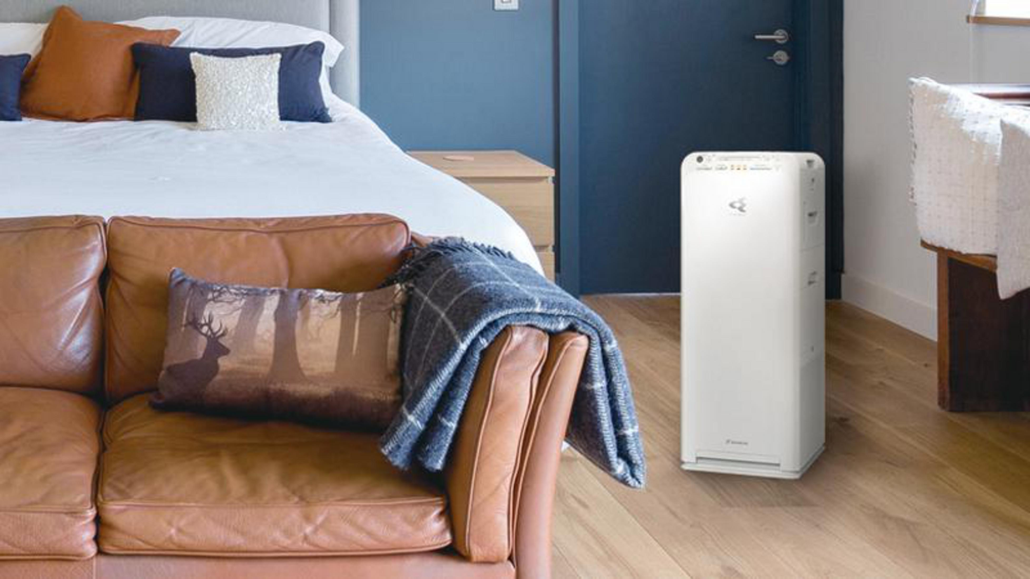 Un divano in pelle con coperta e cuscini davanti a un letto, accanto a un purificatore d'aria bianco.