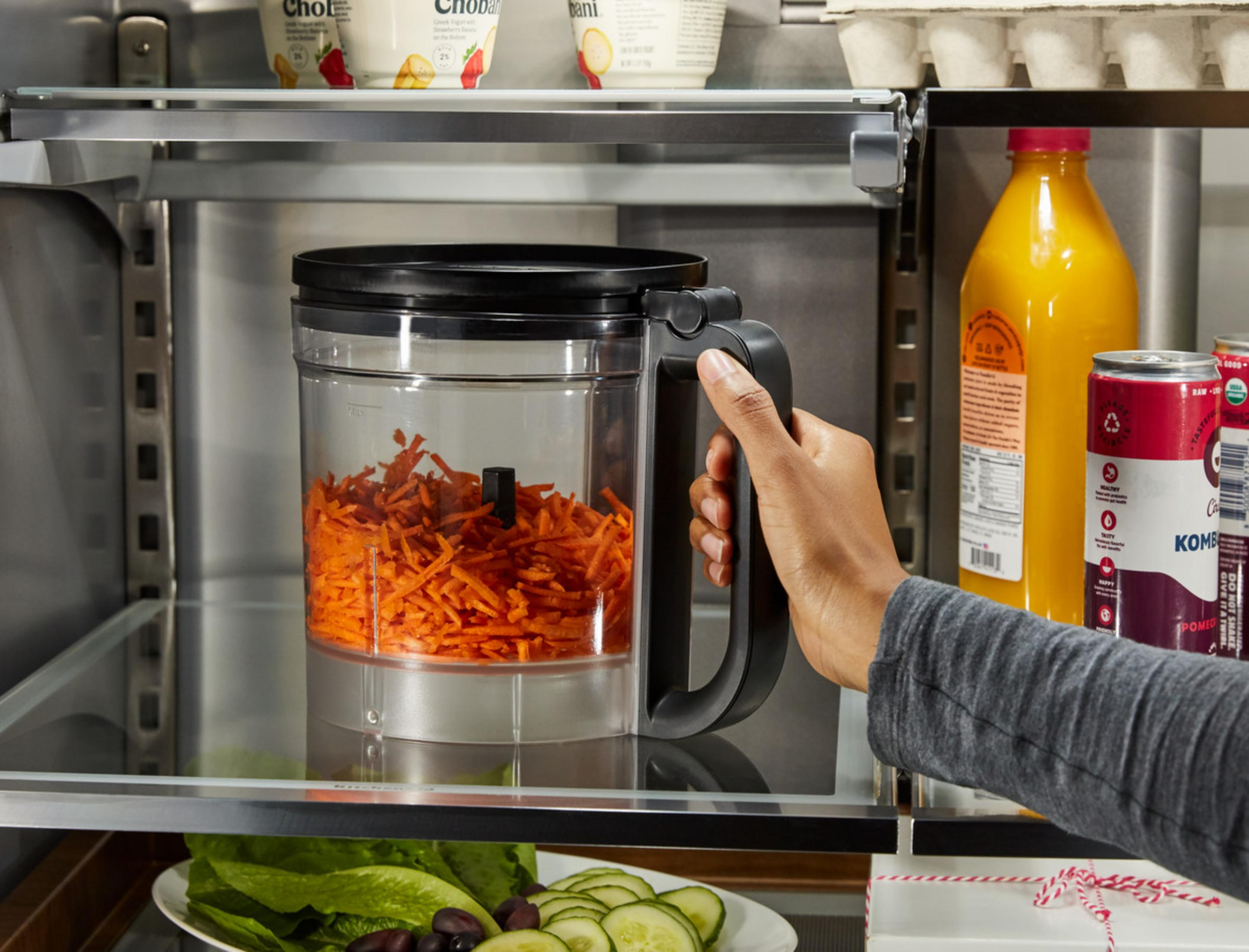 Una mano afferra un robot da cucina con carote grattugiate. Succo d'arancia e kombucha visibili.