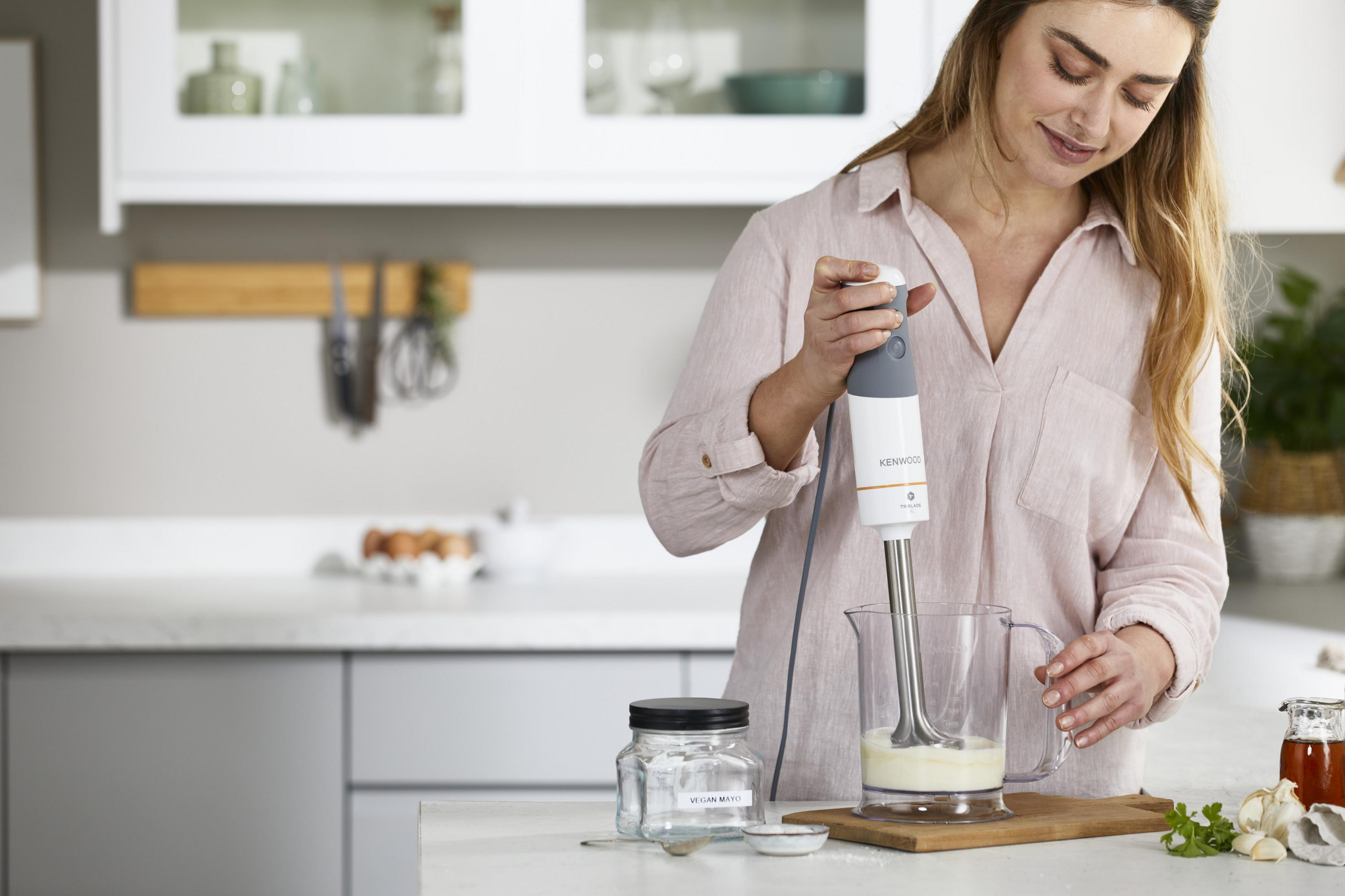 Kobieta miksuje biały płyn w dzbanku blenderem ręcznym w kuchni.