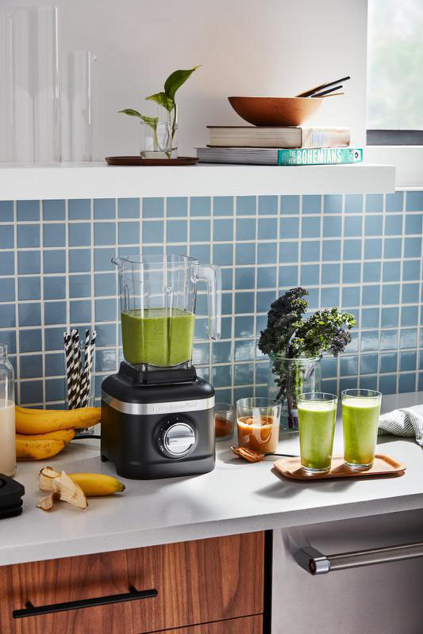 Eine schwarze Küchenmaschine mit grünem Smoothie, Bananen und Smoothie-Gläsern.