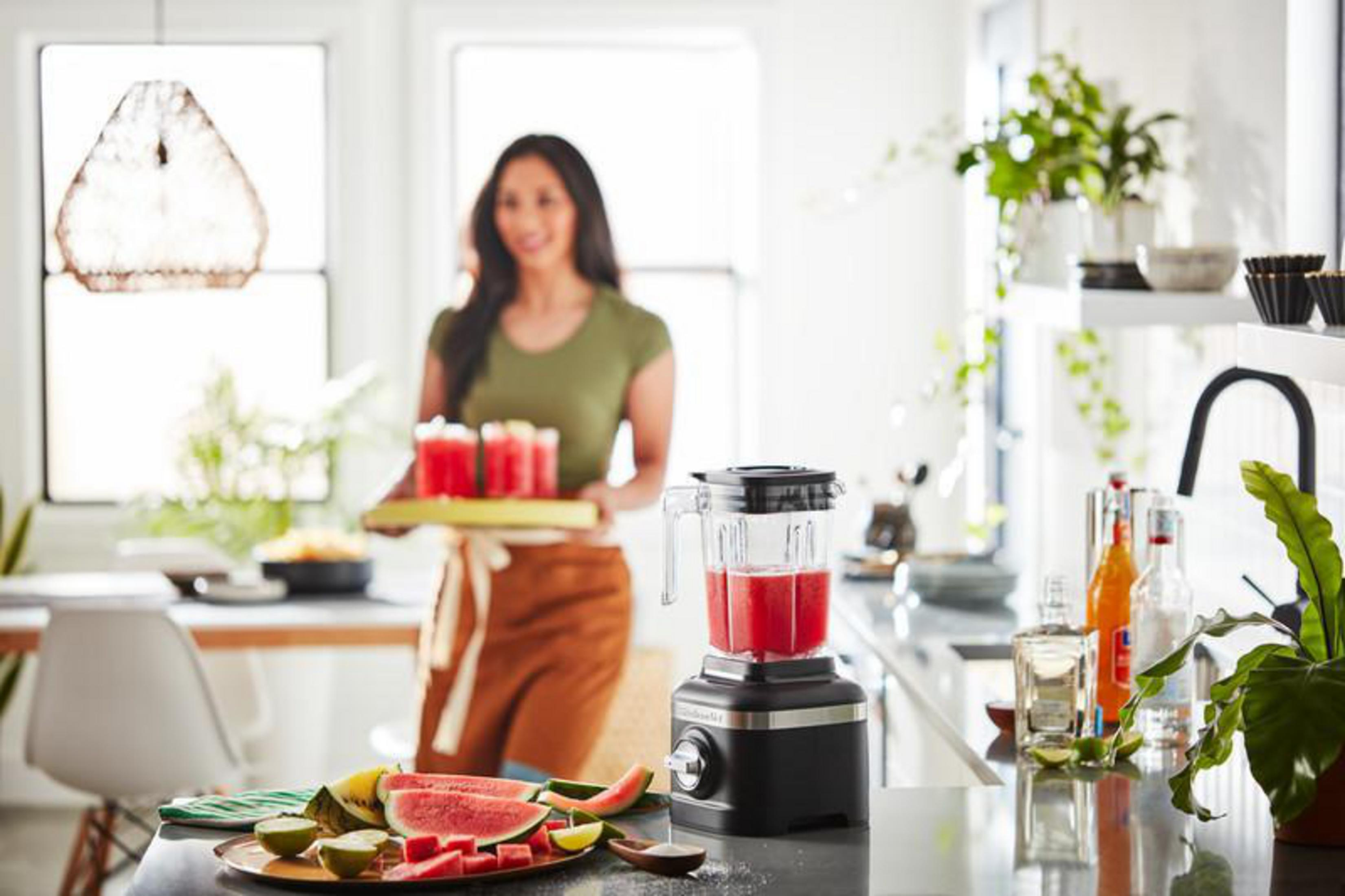 Eine Frau bringt eine Platte mit Gläsern, ein Mixer steht bereit.