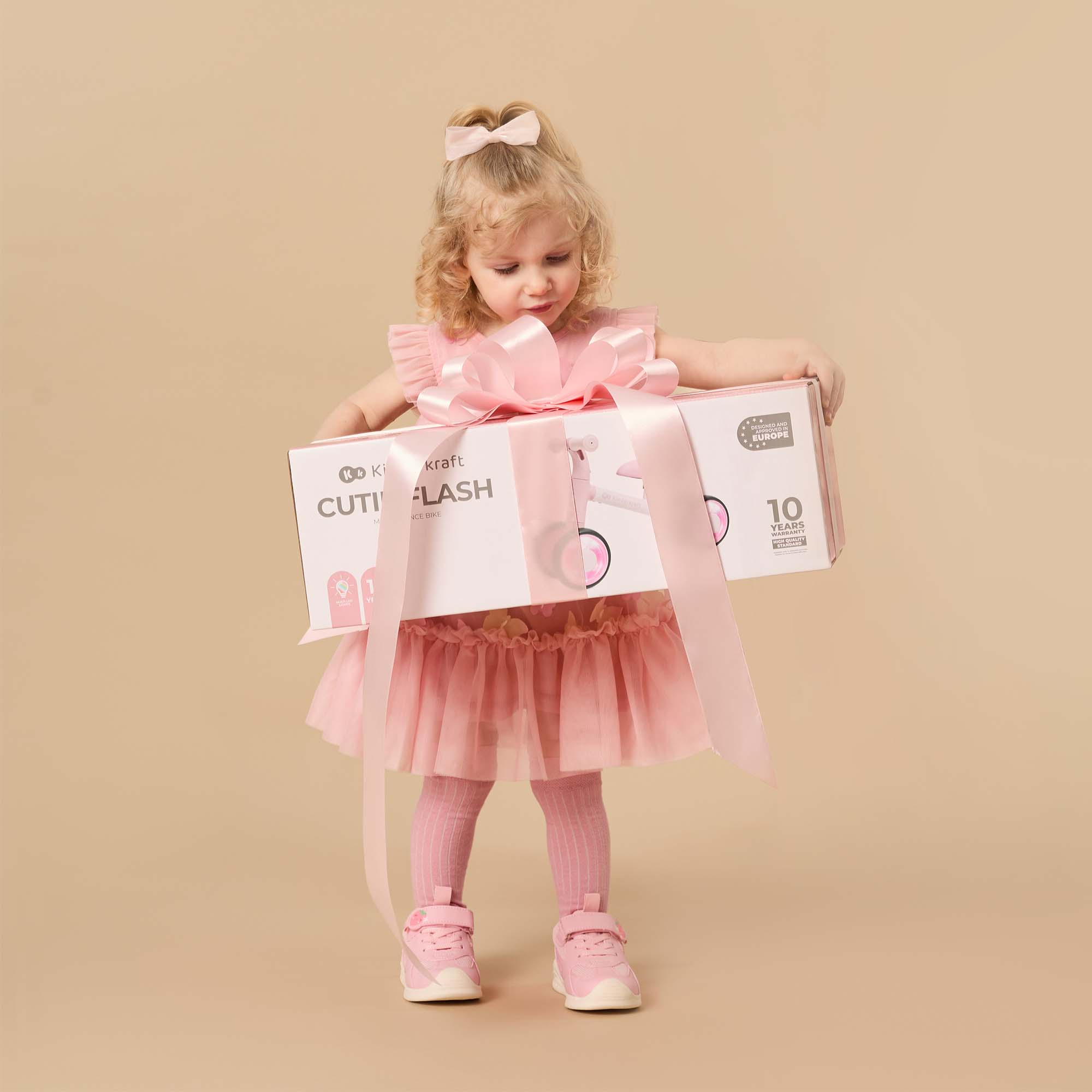Ein kleines Mädchen im rosa Kleid hält eine Geschenkbox mit einem Laufrad.