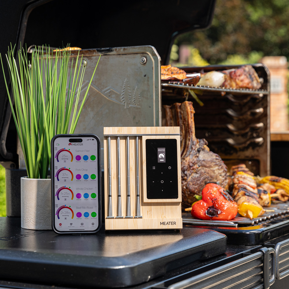 Un ensemble de thermomètres de grill avec application et viande grillée. Une plante en pot est à côté.
