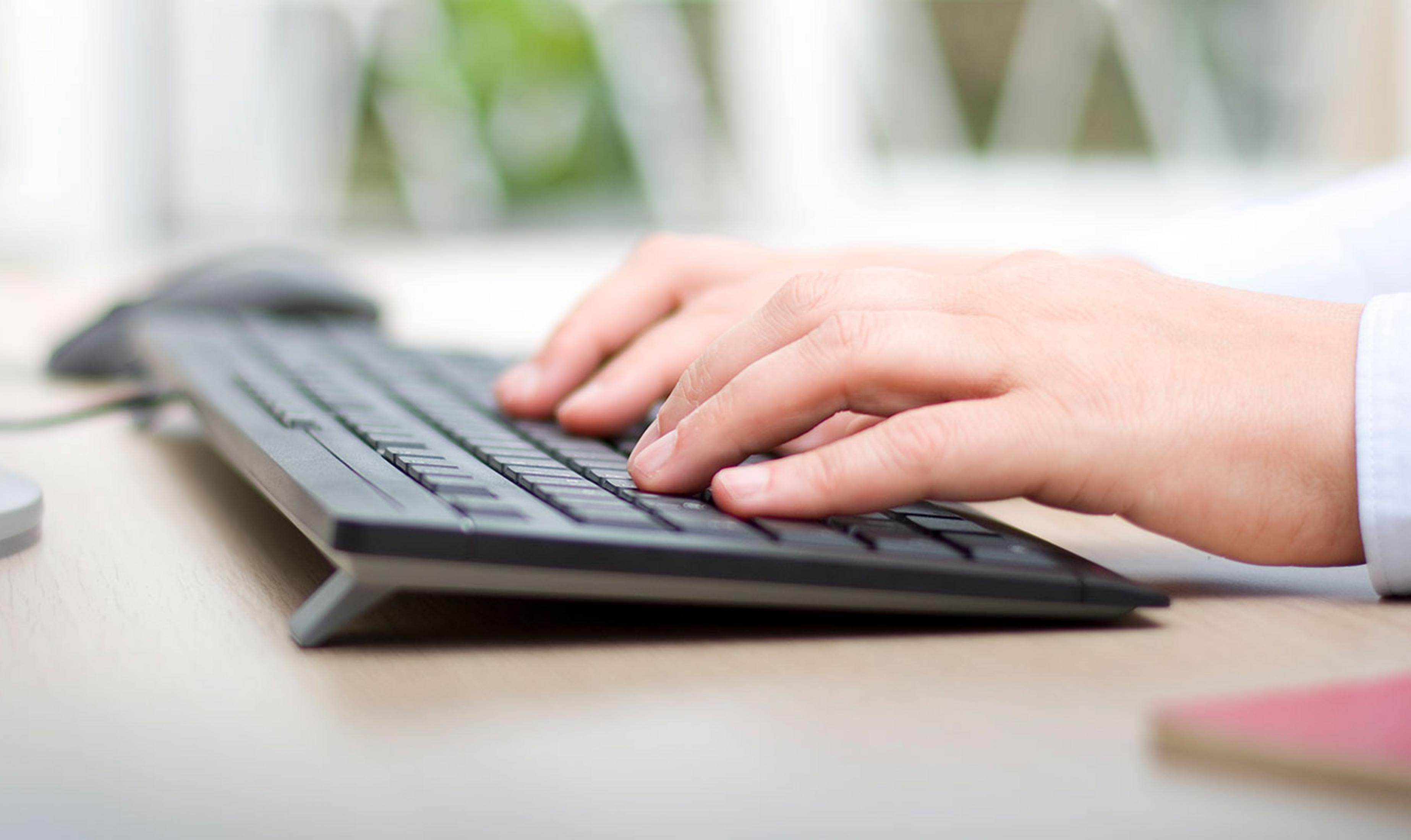 Manos escribiendo en un teclado sobre un escritorio de madera. Un ratón al fondo.