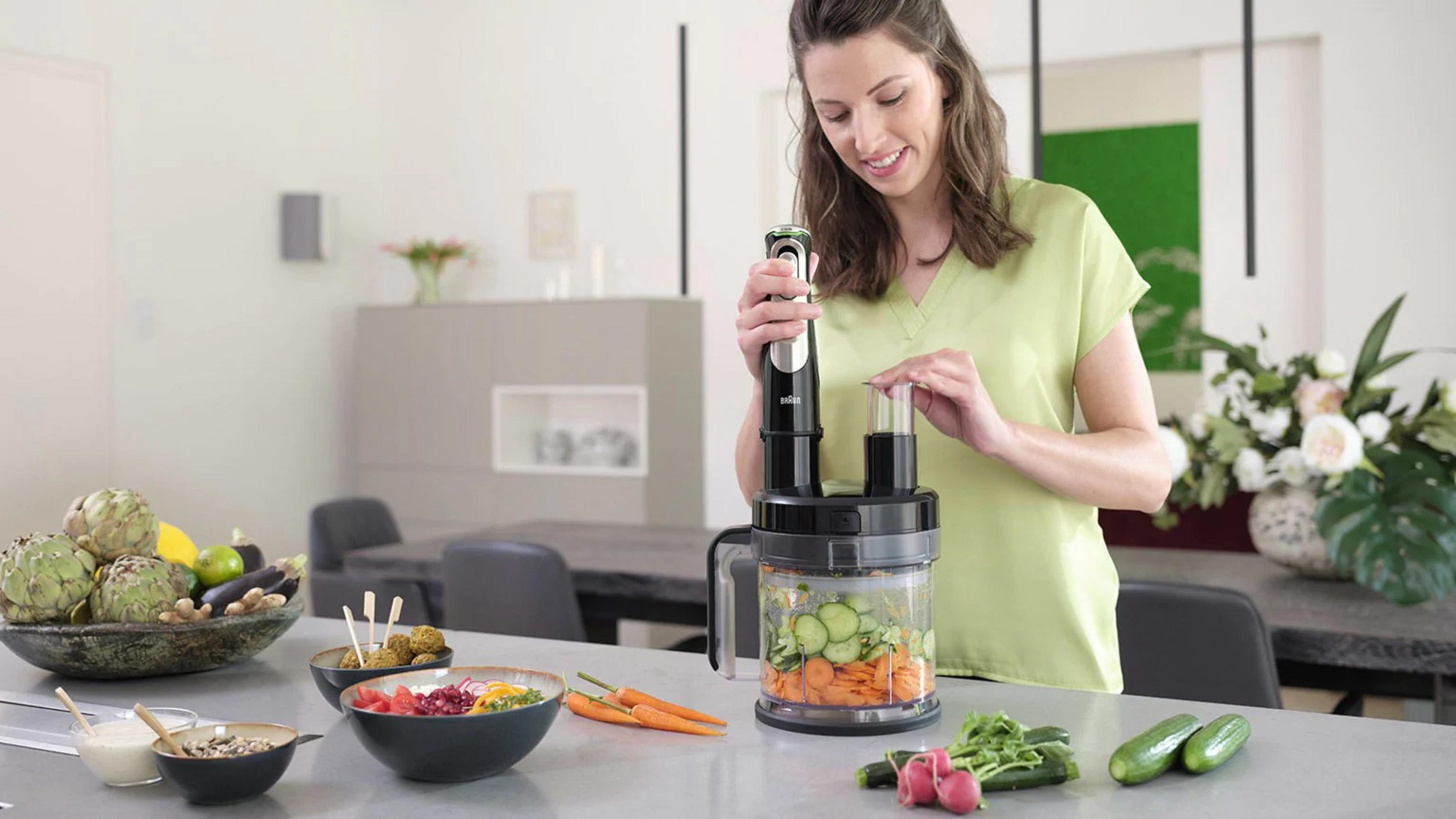 Kobieta używa robota kuchennego w kuchni obok misek z warzywami i falafelem.