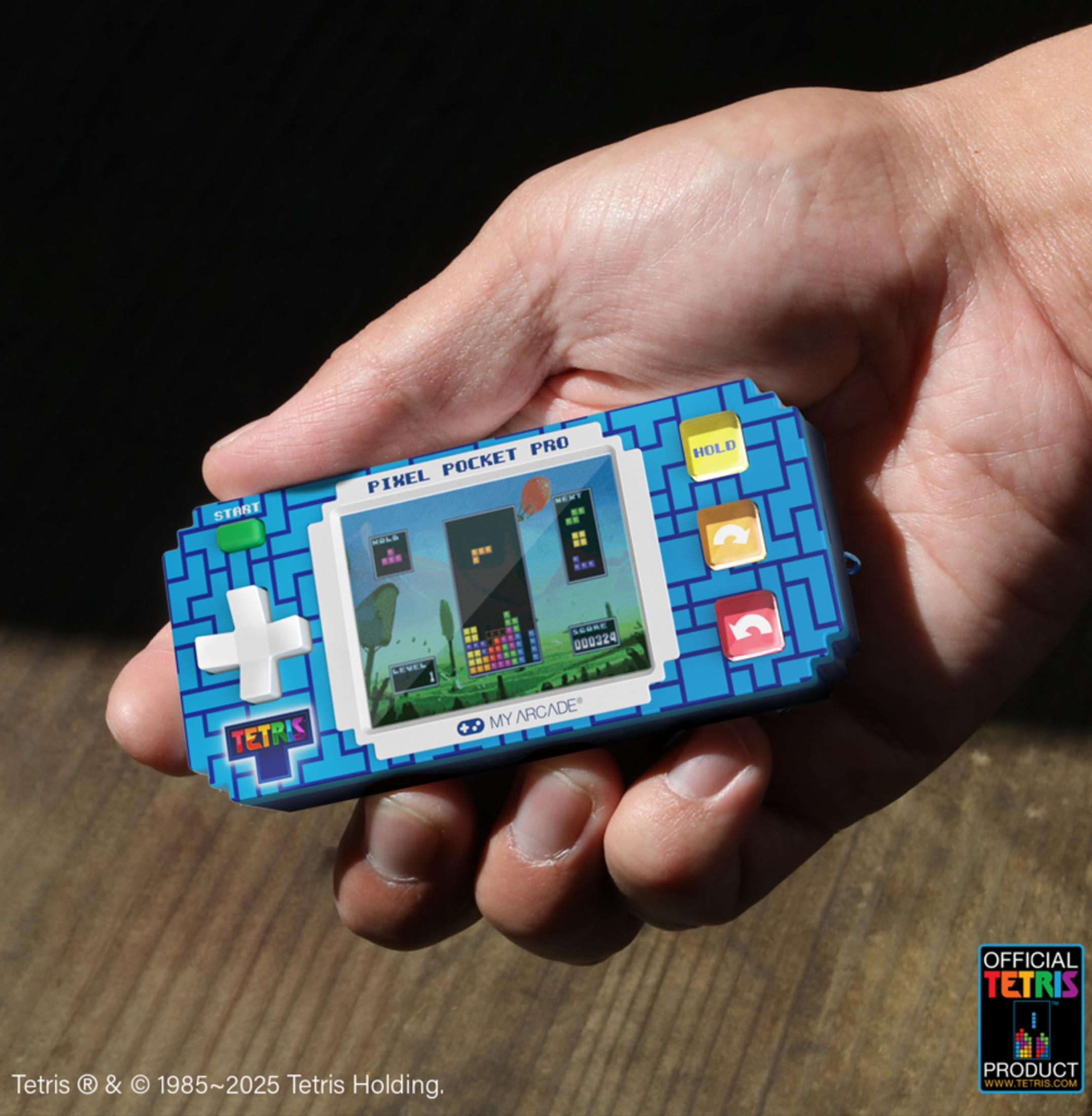 Una mano tiene una console portatile blu Tetris con un gioco Tetris in pixel art.