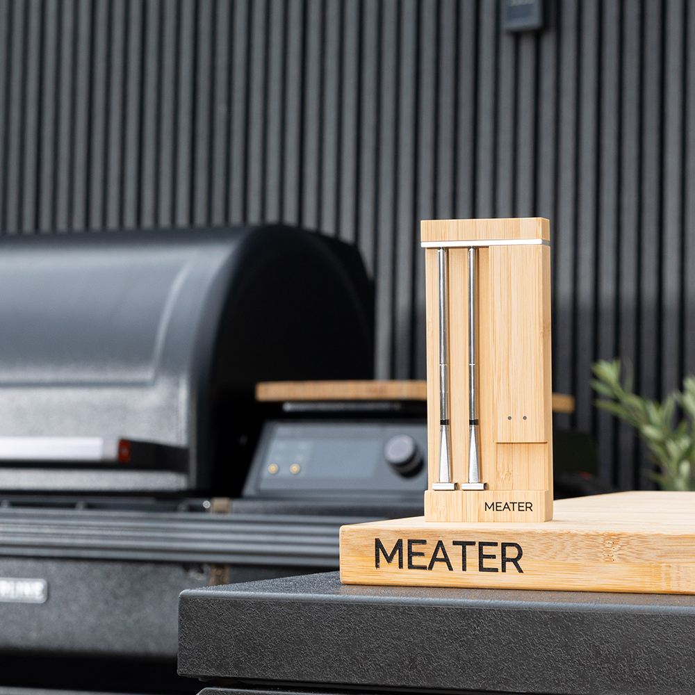 Un support de thermomètre de cuisine en bois avec deux sondes est placé devant un gril.