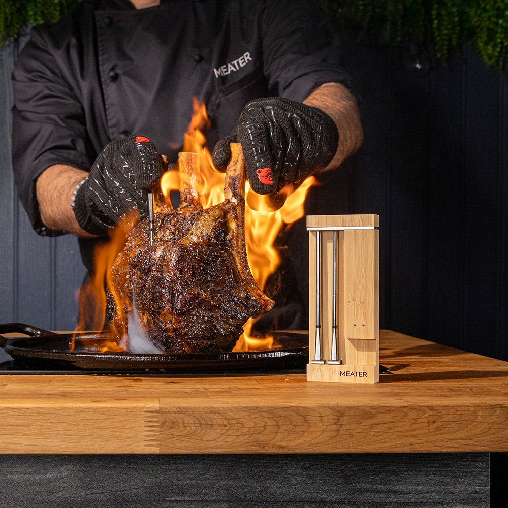Chef en manteau noir cuit un rôti de côte première sur flammes vives, avec un support thermomètre MEATER.