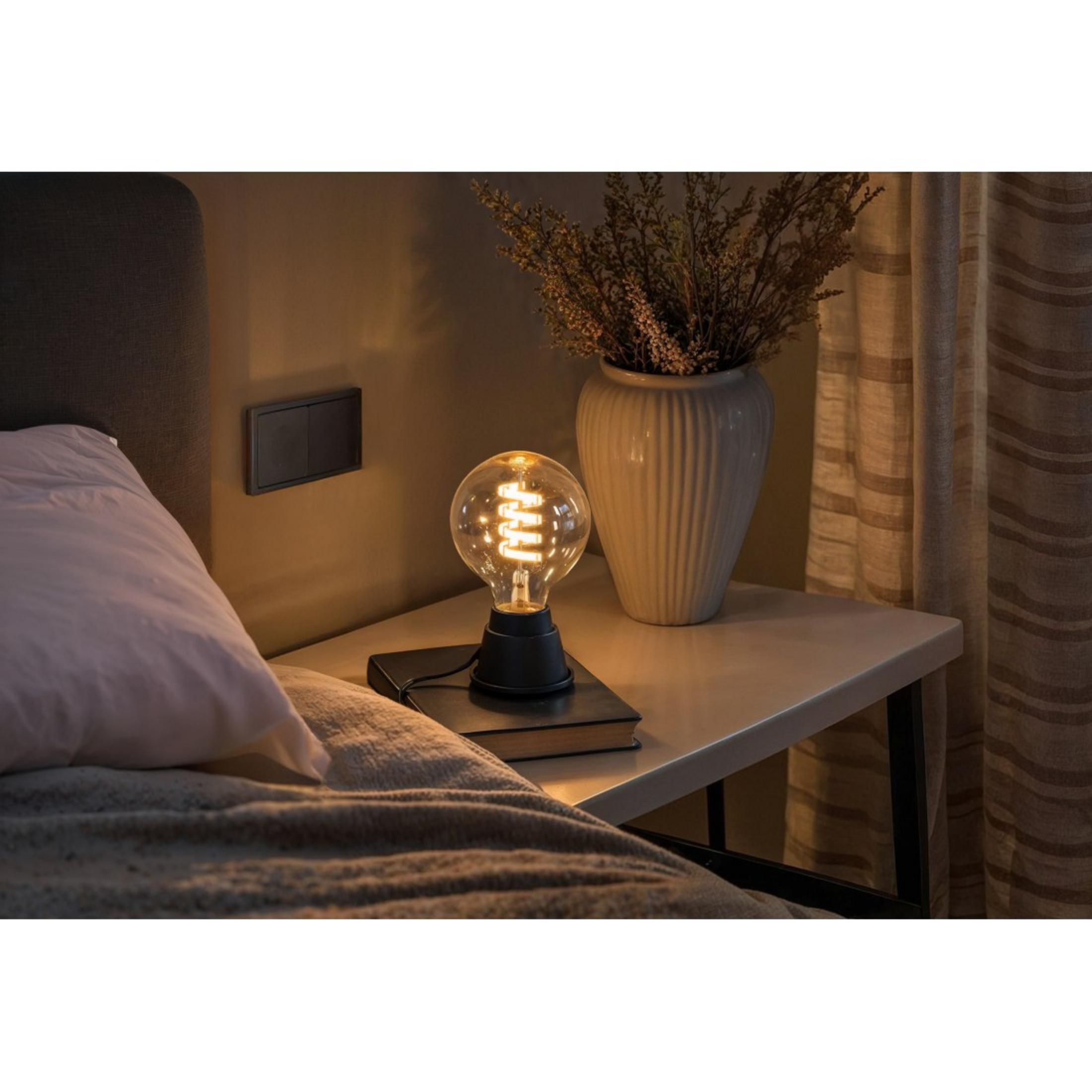 Una lampada da comodino con lampadina a filamento brilla accanto a un letto con cuscini.