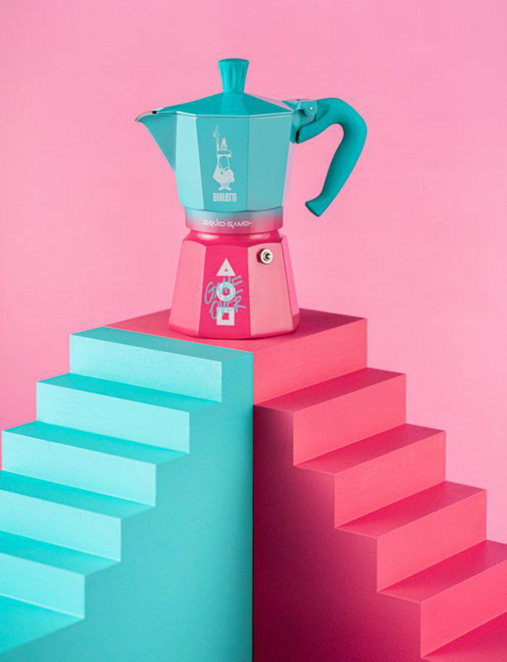Turkusowa kawiarka Moka stoi na różowo-turkusowych schodach na różowym tle.