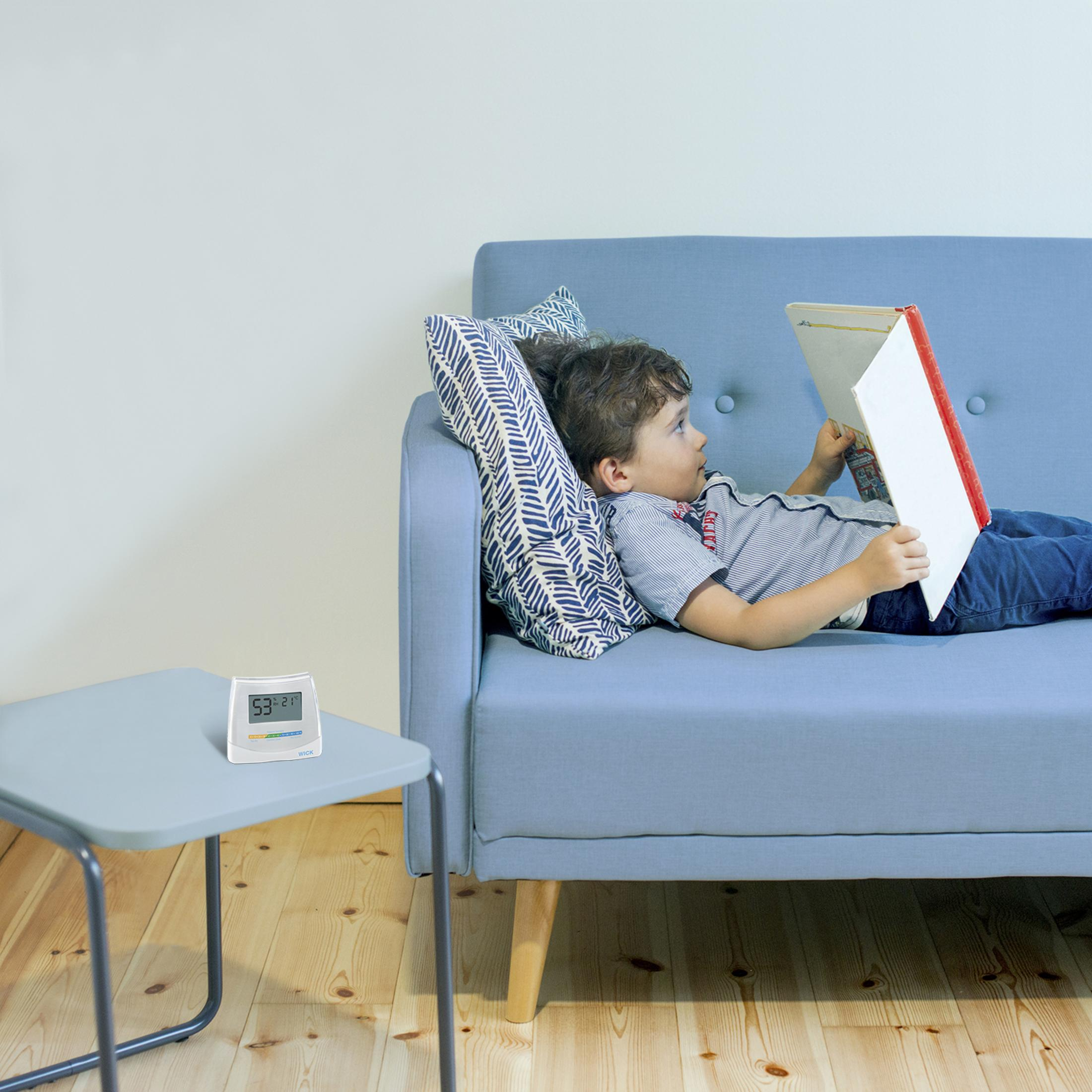 Ein Junge liegt auf einem Sofa und liest ein Buch. Daneben steht ein Tisch mit einem Thermometer.