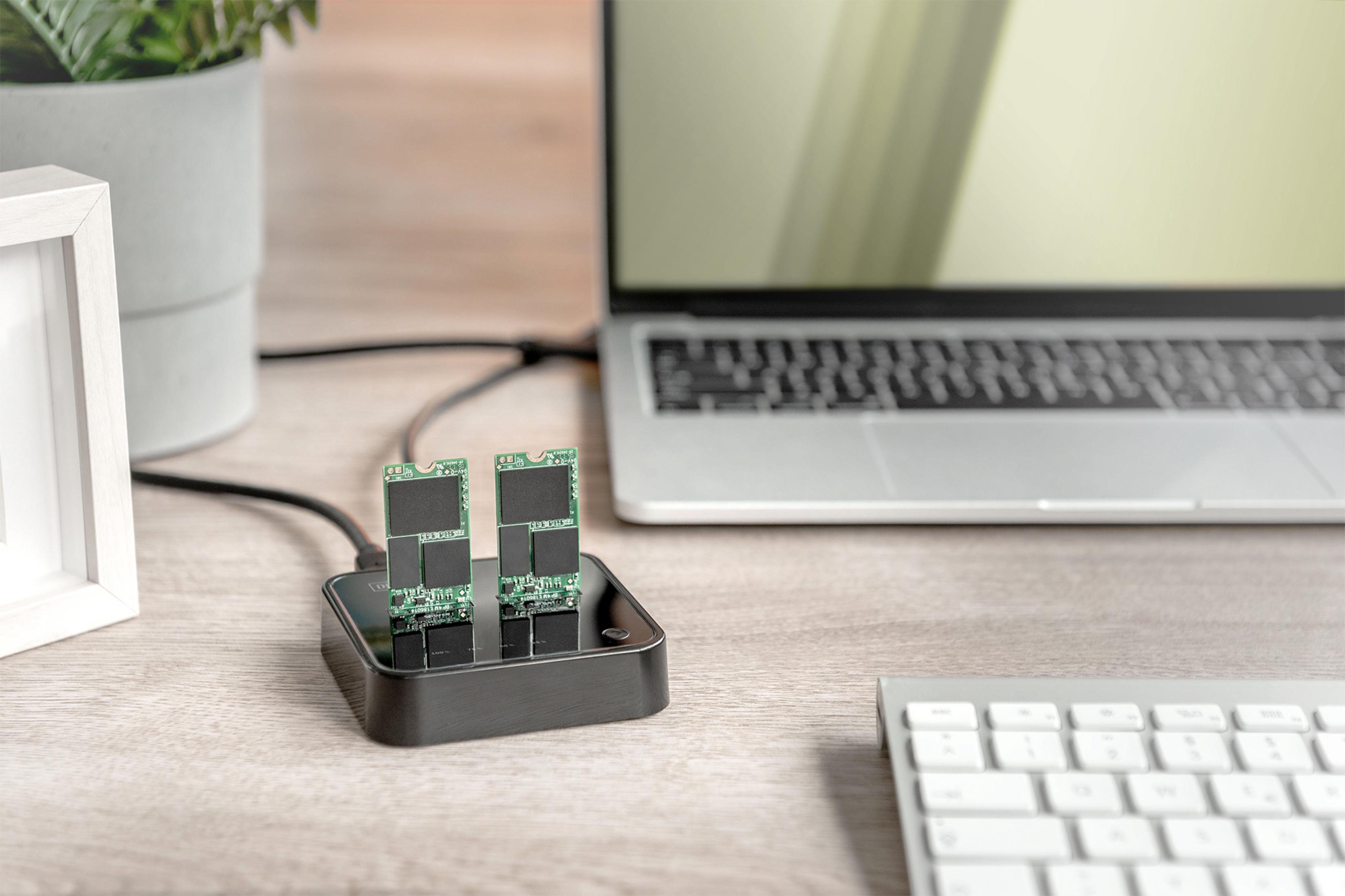 Twee SSD's op een zwart dockingstation, laptop en toetsenbord op de achtergrond.
