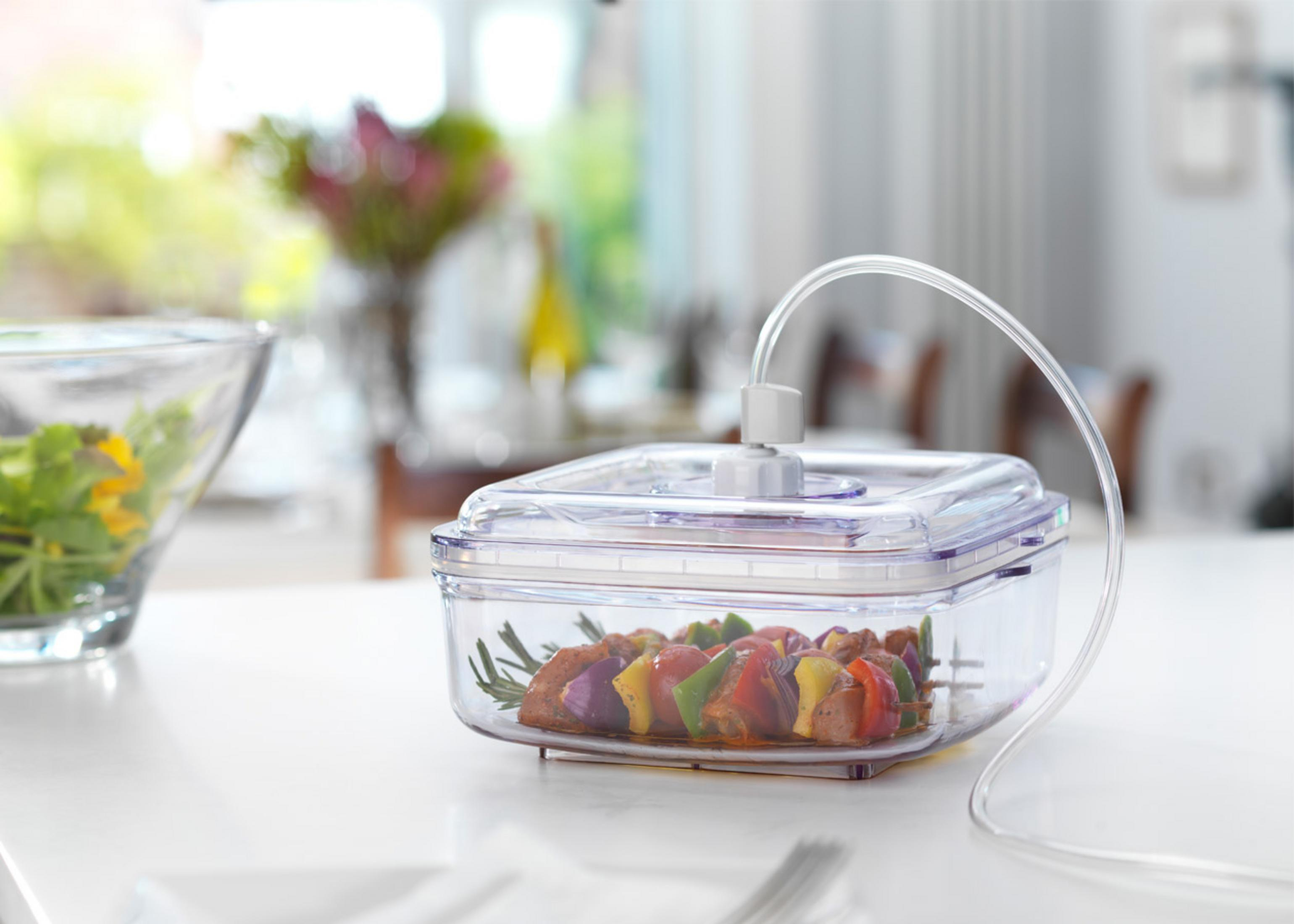 Un recipiente al vacío con brochetas de carne y hierbas junto a una ensalada.