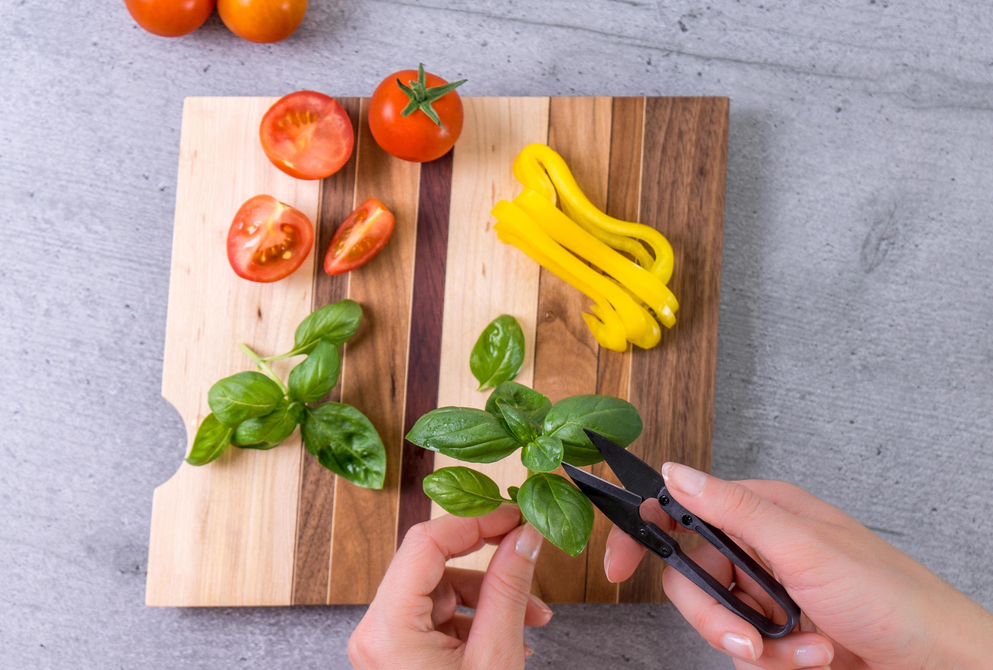 Mani che tagliano basilico su tagliere in legno con pomodori e peperoni gialli.