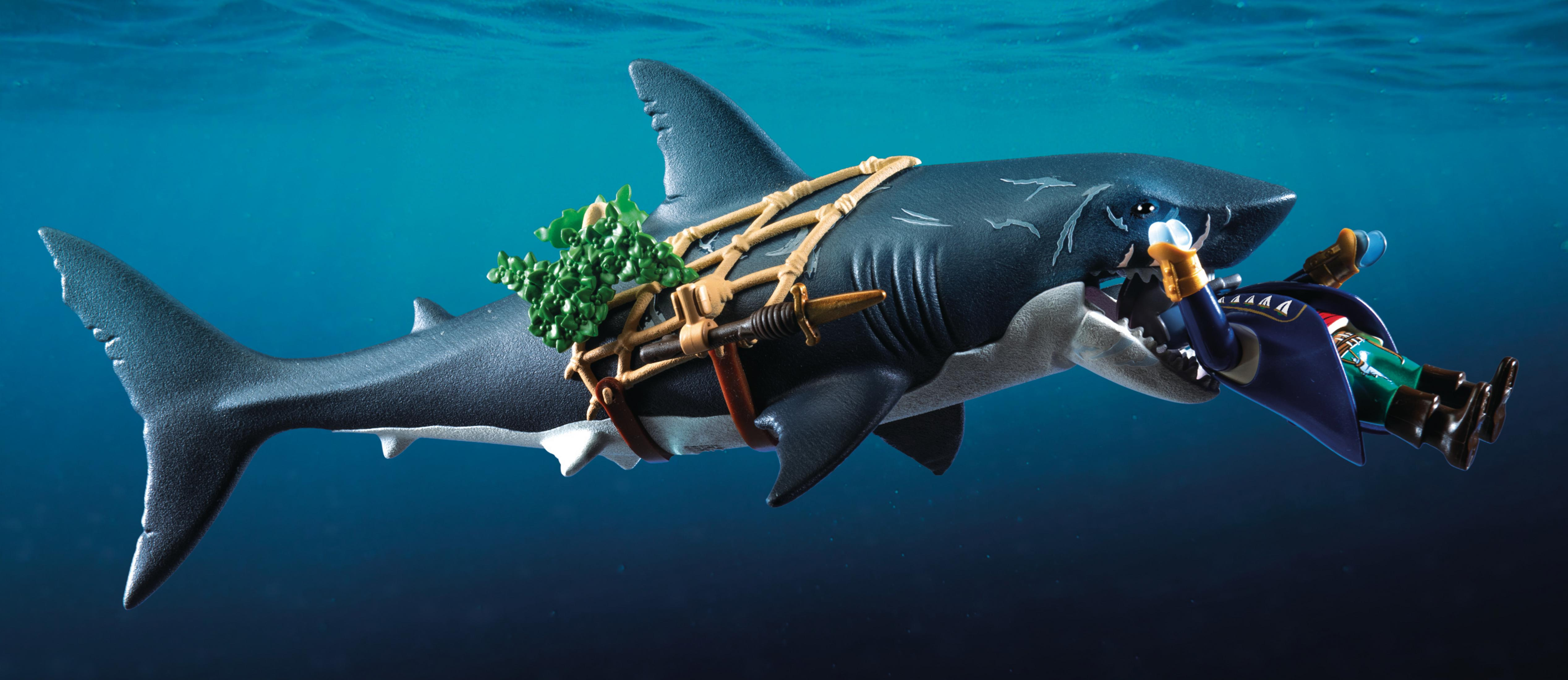 Zabawkowy rekin z piratem w pysku pływa pod wodą. Przyczepione są zielone rośliny.