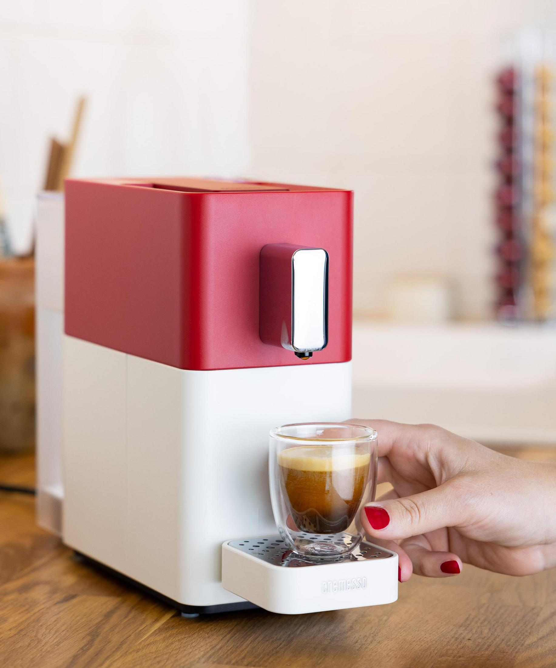 Eine rote und weiße Kaffeemaschine brüht Espresso in ein Glas, das eine Hand hält.