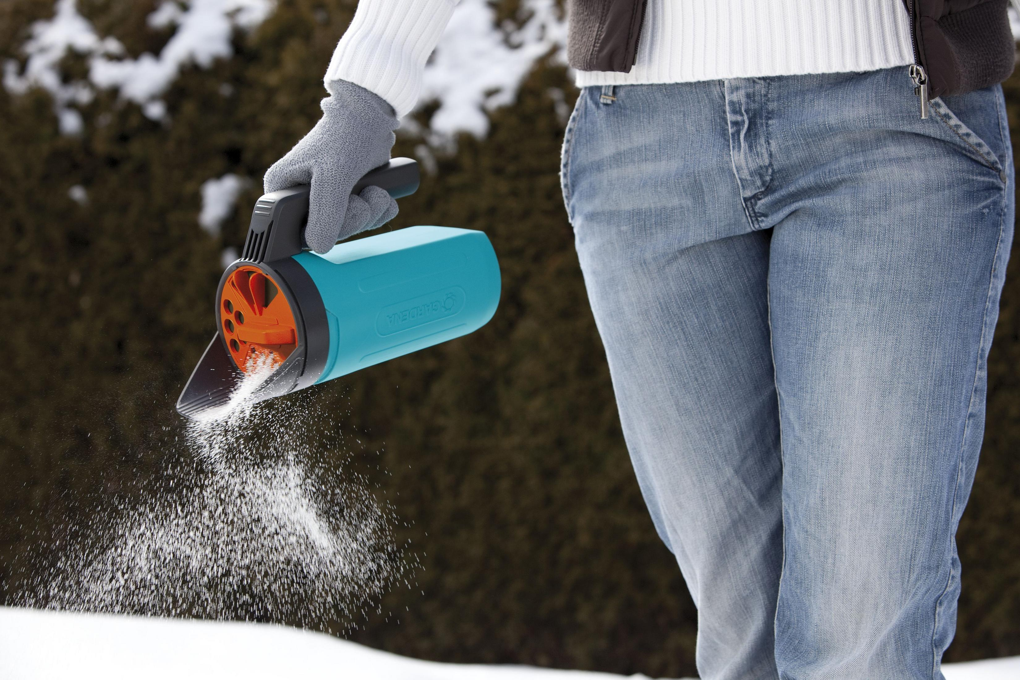 Ręka w szarej rękawiczce trzyma niebieski rozsiewacz, rozsypując sól na śniegu.