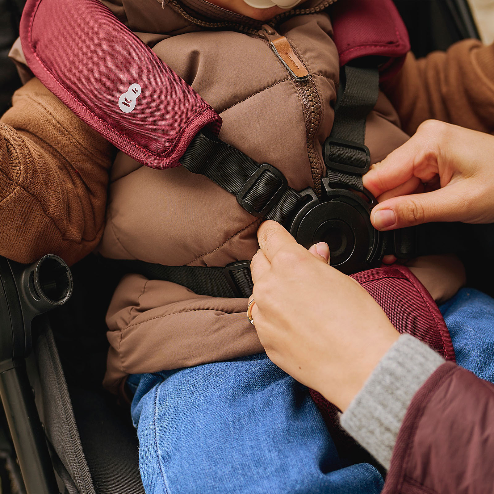 Hände schließen einen Kinderwagen-Gurt über einem Kind in braunem Wintermantel.