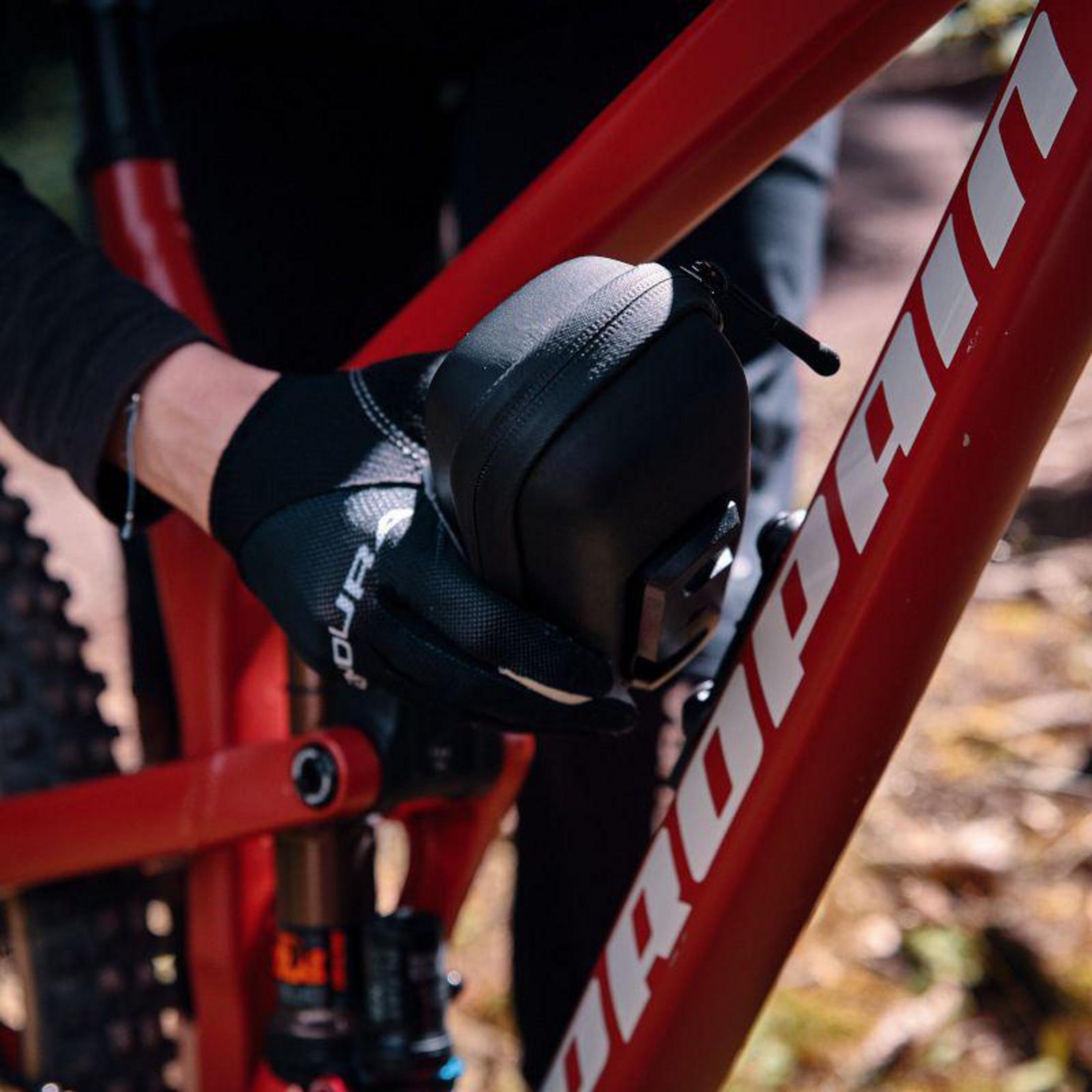 Osoba w czarnych rękawiczkach trzyma czarną torbę na czerwonej ramie roweru.