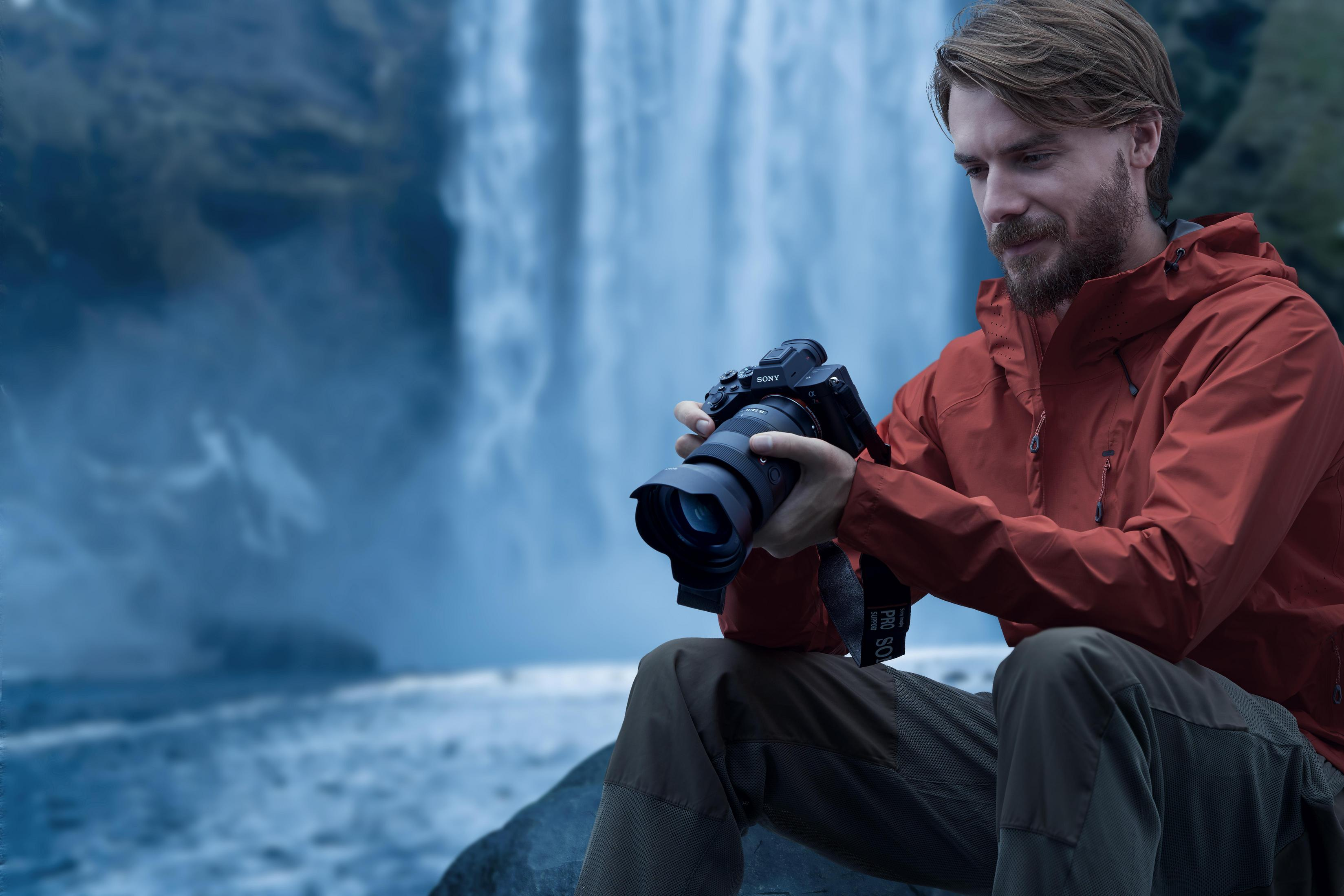 Man with beard in orange jacket holds camera in front of waterfall.