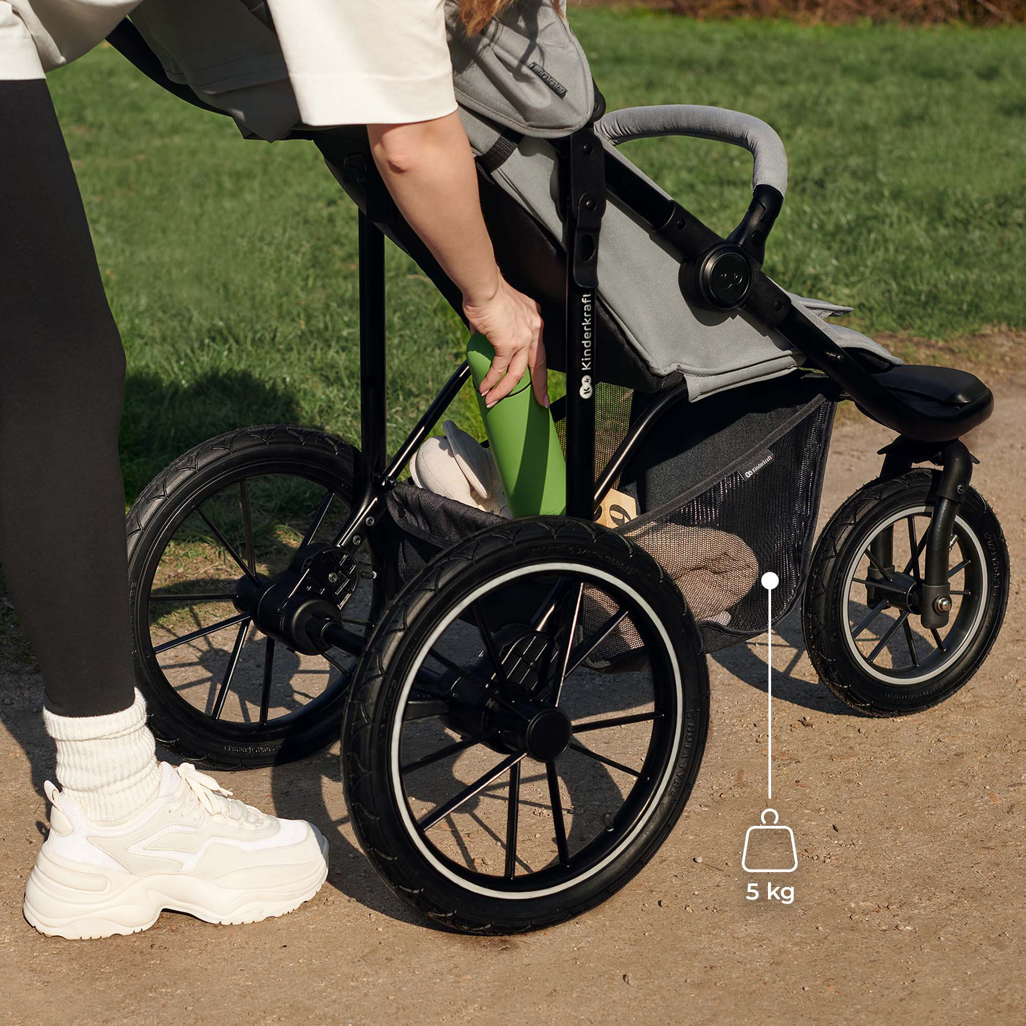 Eine Person verstaut eine grüne Flasche in einem Kinderwagen. Der Kinderwagen hat große Räder.