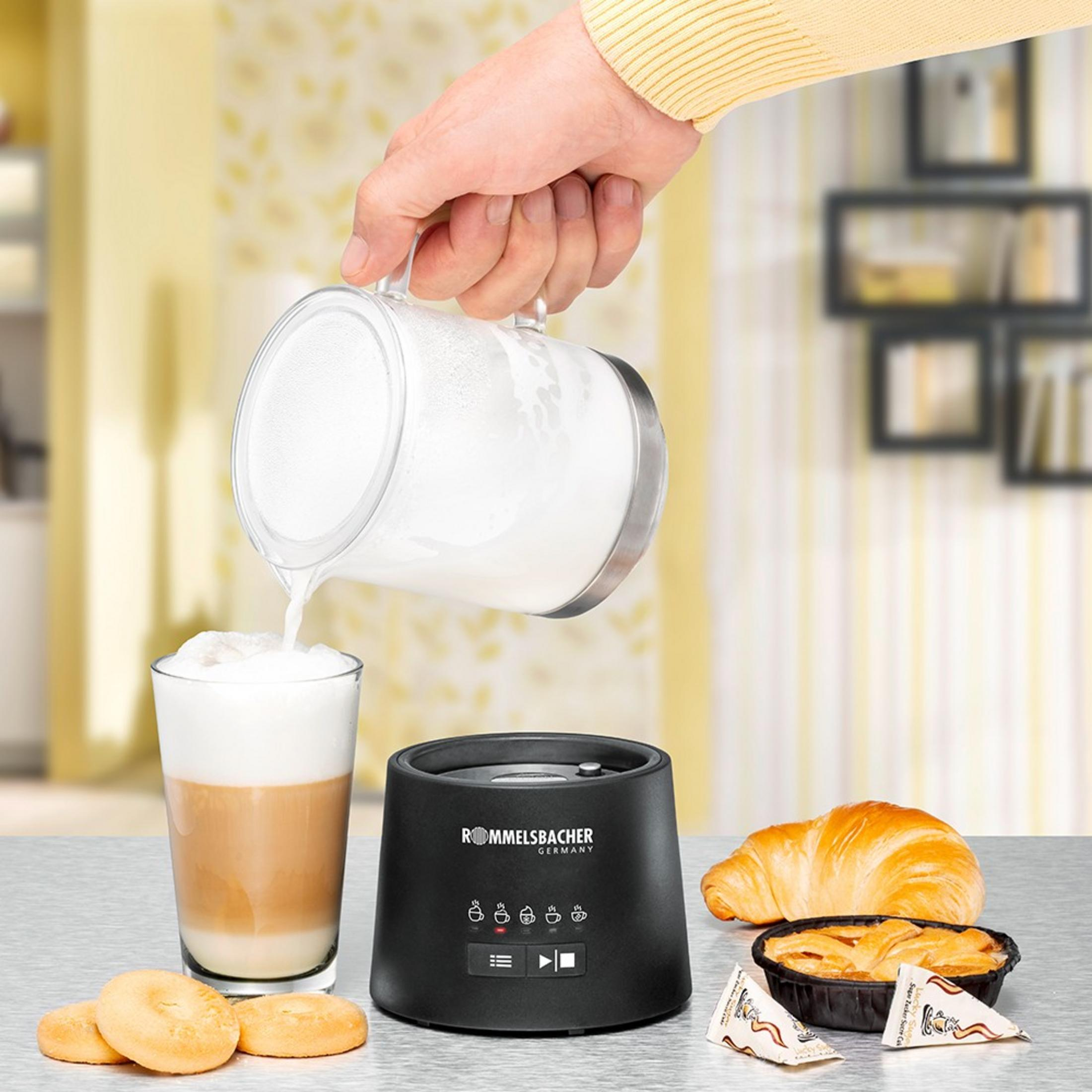 Schaum wird aus einem Krug in ein Glas Latte gegossen. Daneben ein Milchaufschäumer.