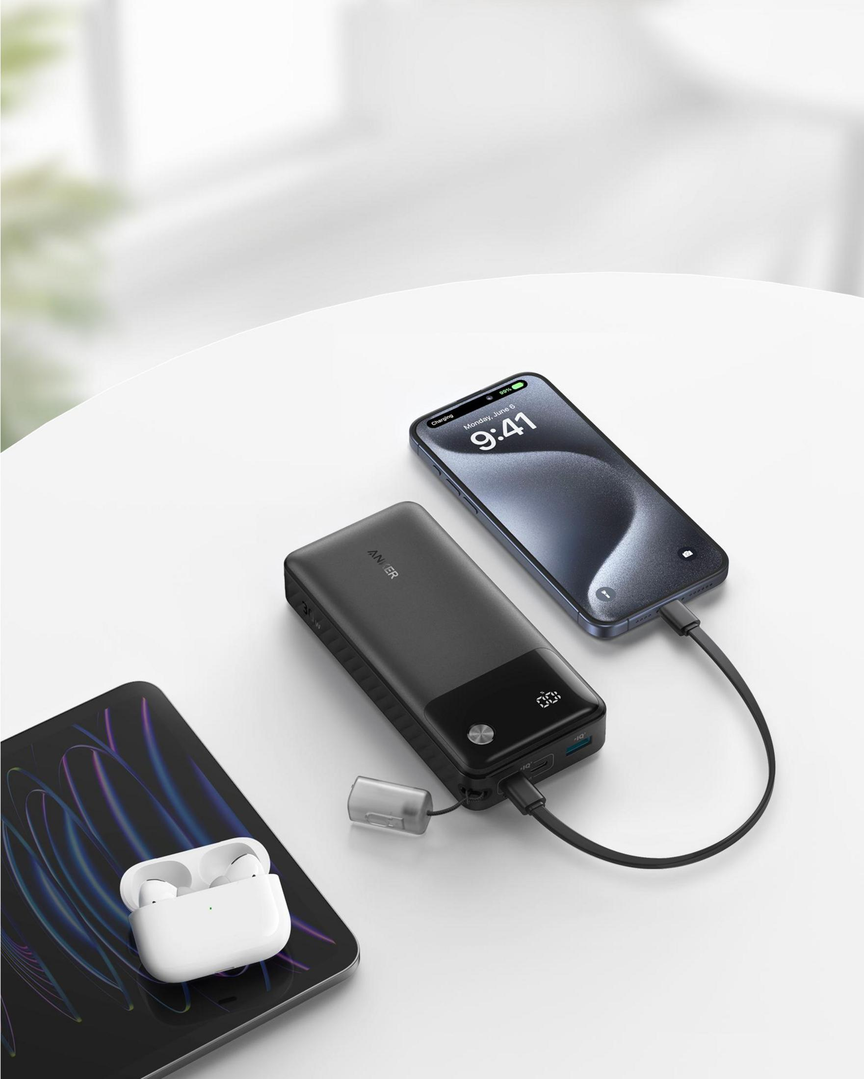 Power bank nera carica un iPhone, con AirPods in custodia su tablet vicino.