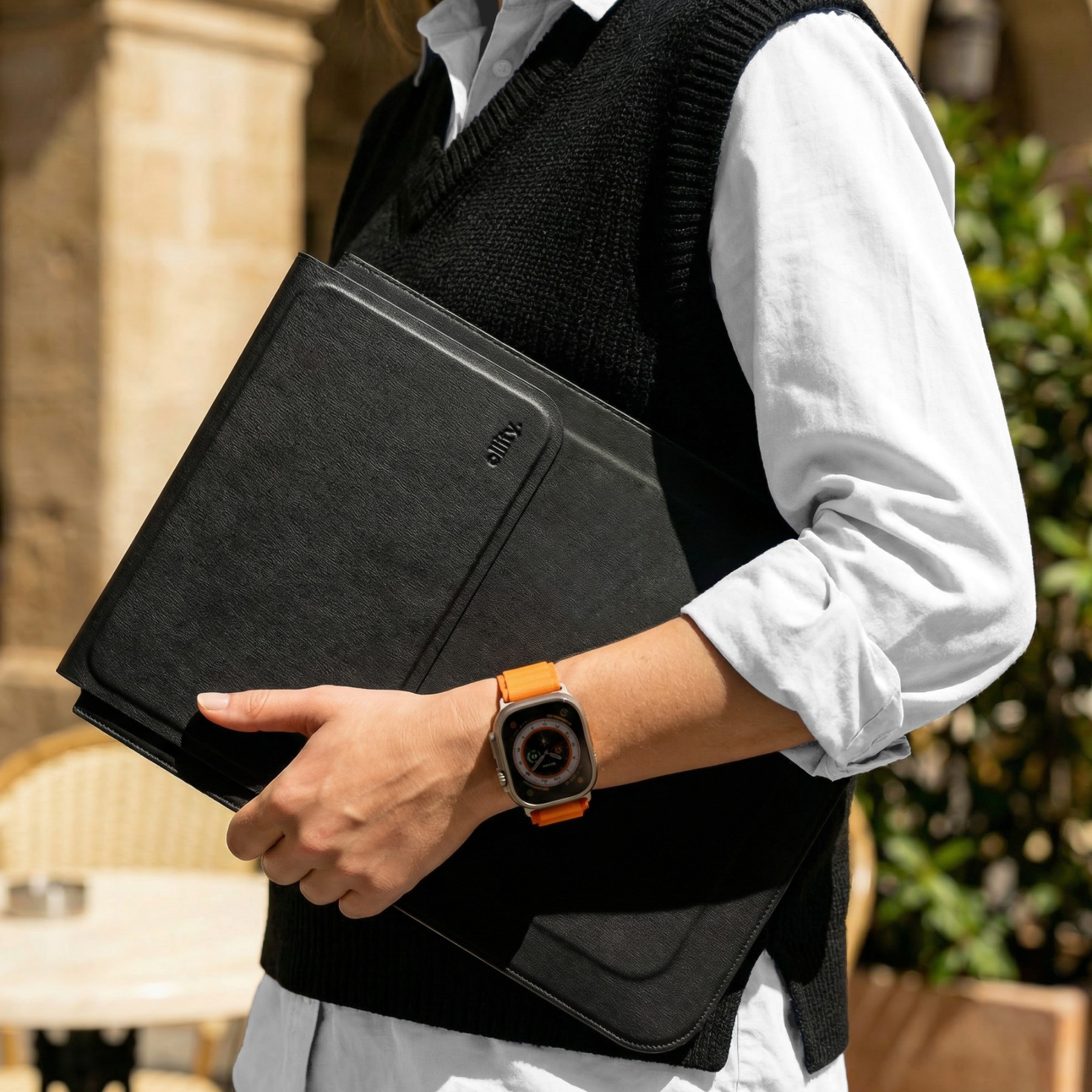 Osoba w czarnej kamizelce i białej koszuli trzyma czarną teczkę. Nosi smartwatch.