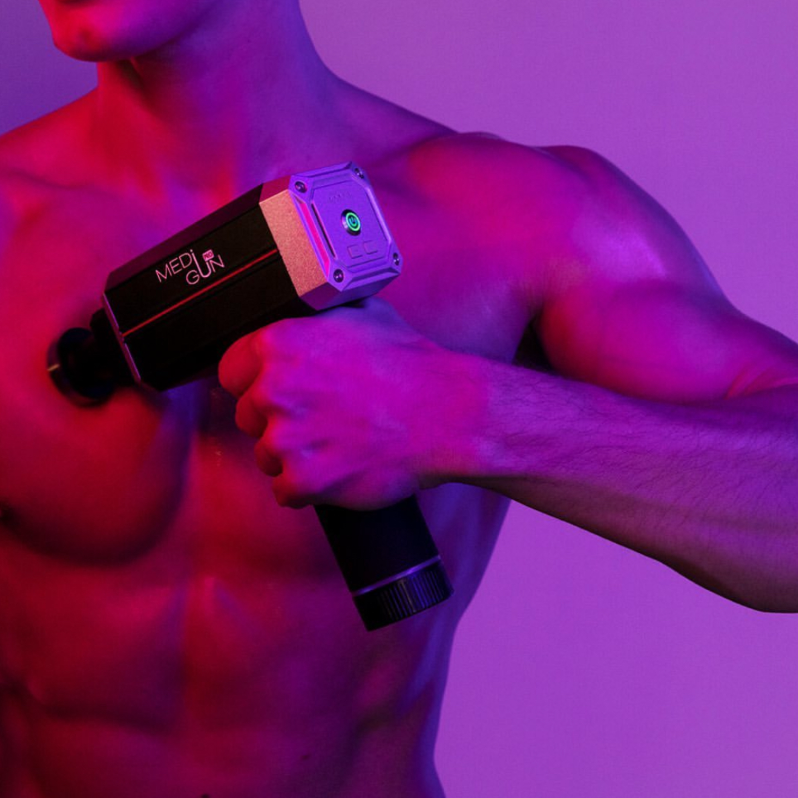 Mężczyzna trzyma czarny pistolet do masażu na piersi, oświetlony różowym i fioletowym światłem.