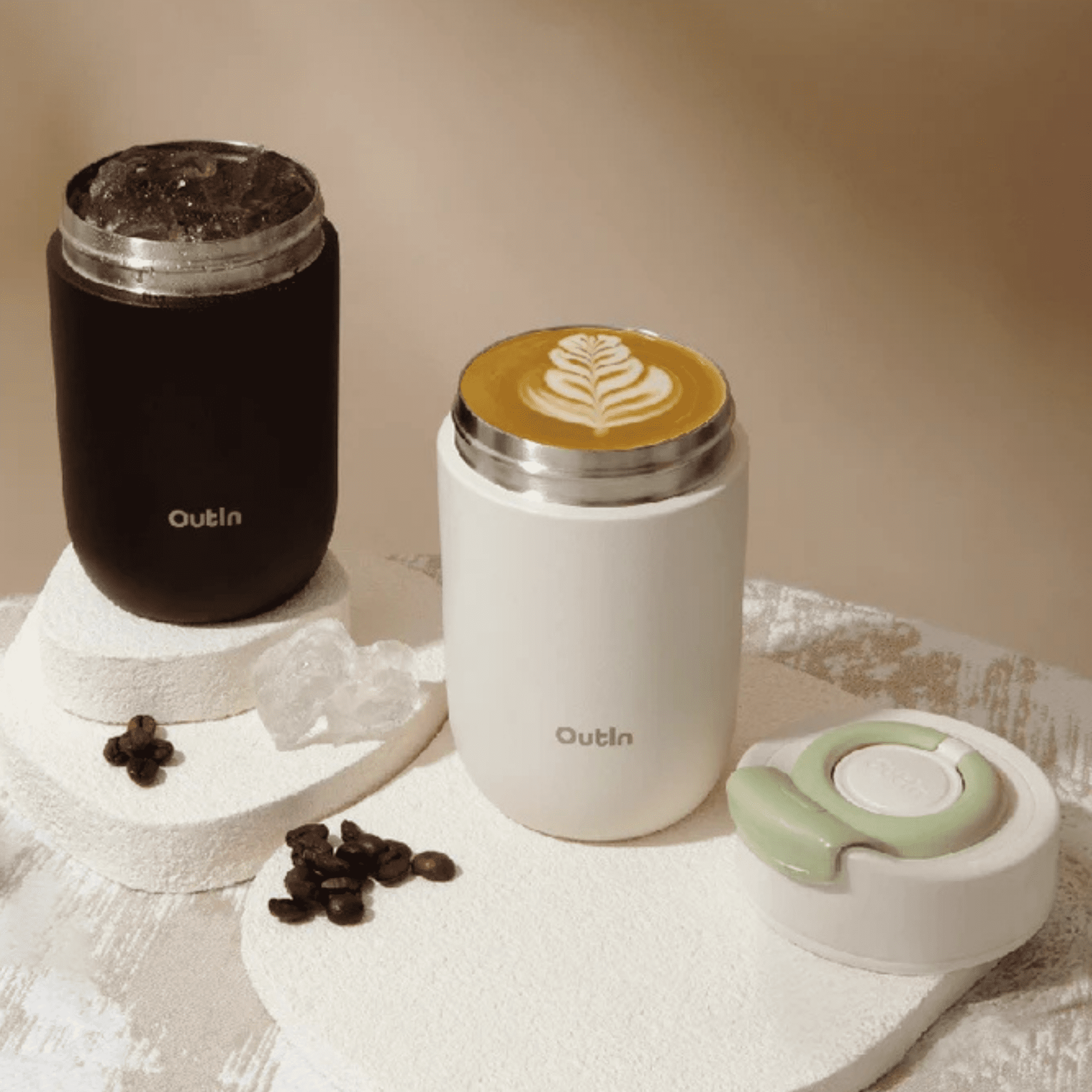 Dwa kubki Outin: czarny z wodą z lodem, biały z latte art. W pobliżu znajdują się ziarna kawy.