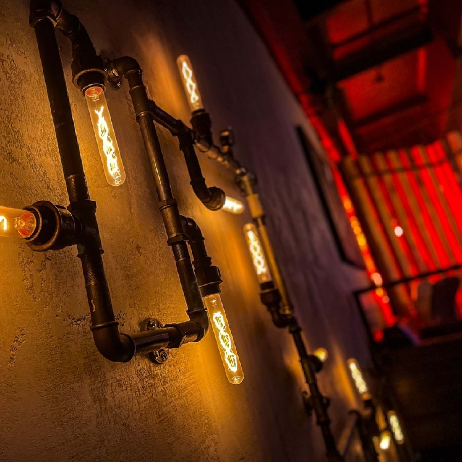 Lámpara de pared industrial con tubos negros y bombillas Edison emitiendo luz cálida.