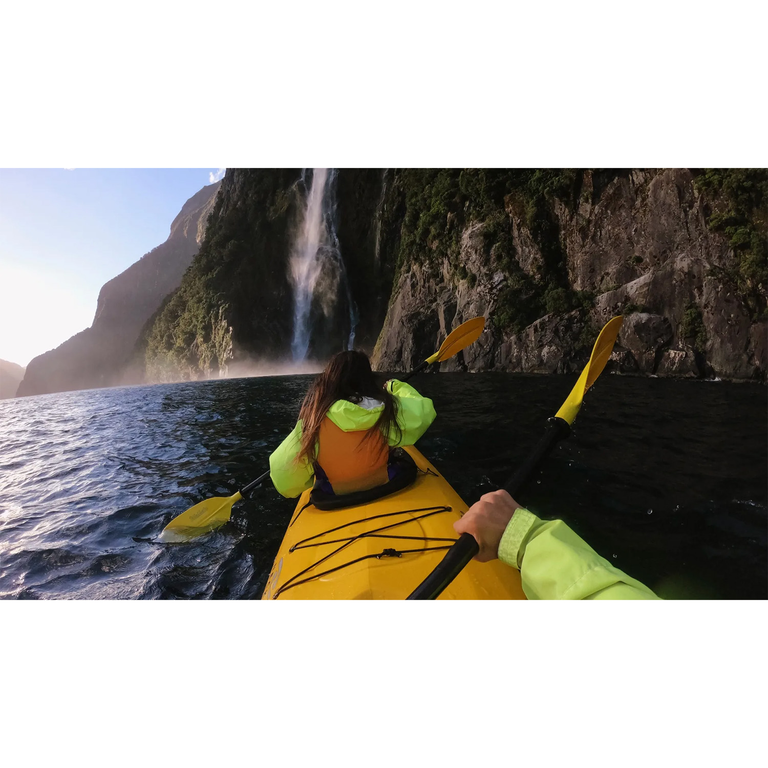 Dwóch kajakarzy płynie łodzią po jeziorze w kierunku wodospadu, otoczeni stromymi, zielonymi górami.