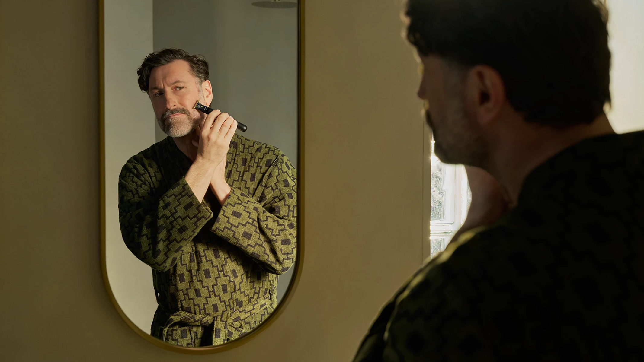 Mann mit Bart rasiert sich im Spiegel mit einem Trimmer in einem grünen Bademantel.