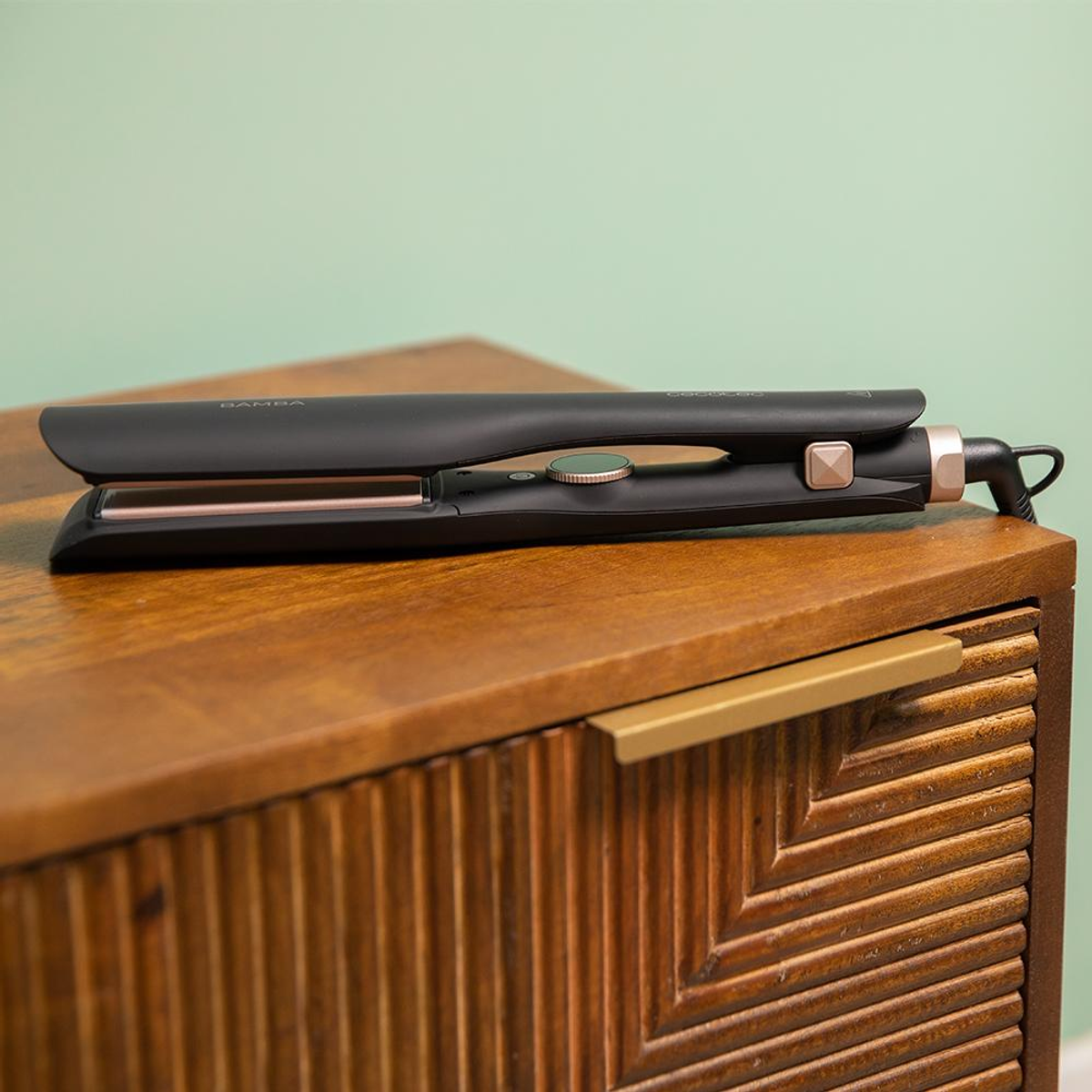 Una piastra per capelli nera poggia su un mobile in legno con sfondo verde.