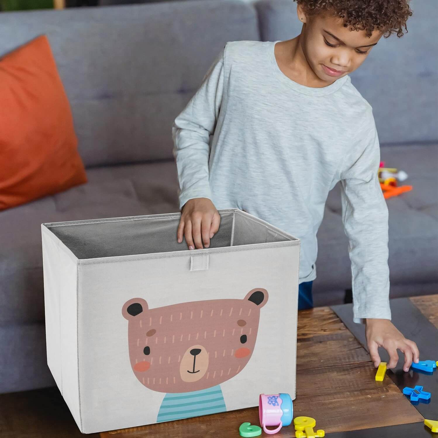 Ein Junge spielt mit Spielzeugbuchstaben neben einer Spielzeugkiste mit Bärenmotiv.