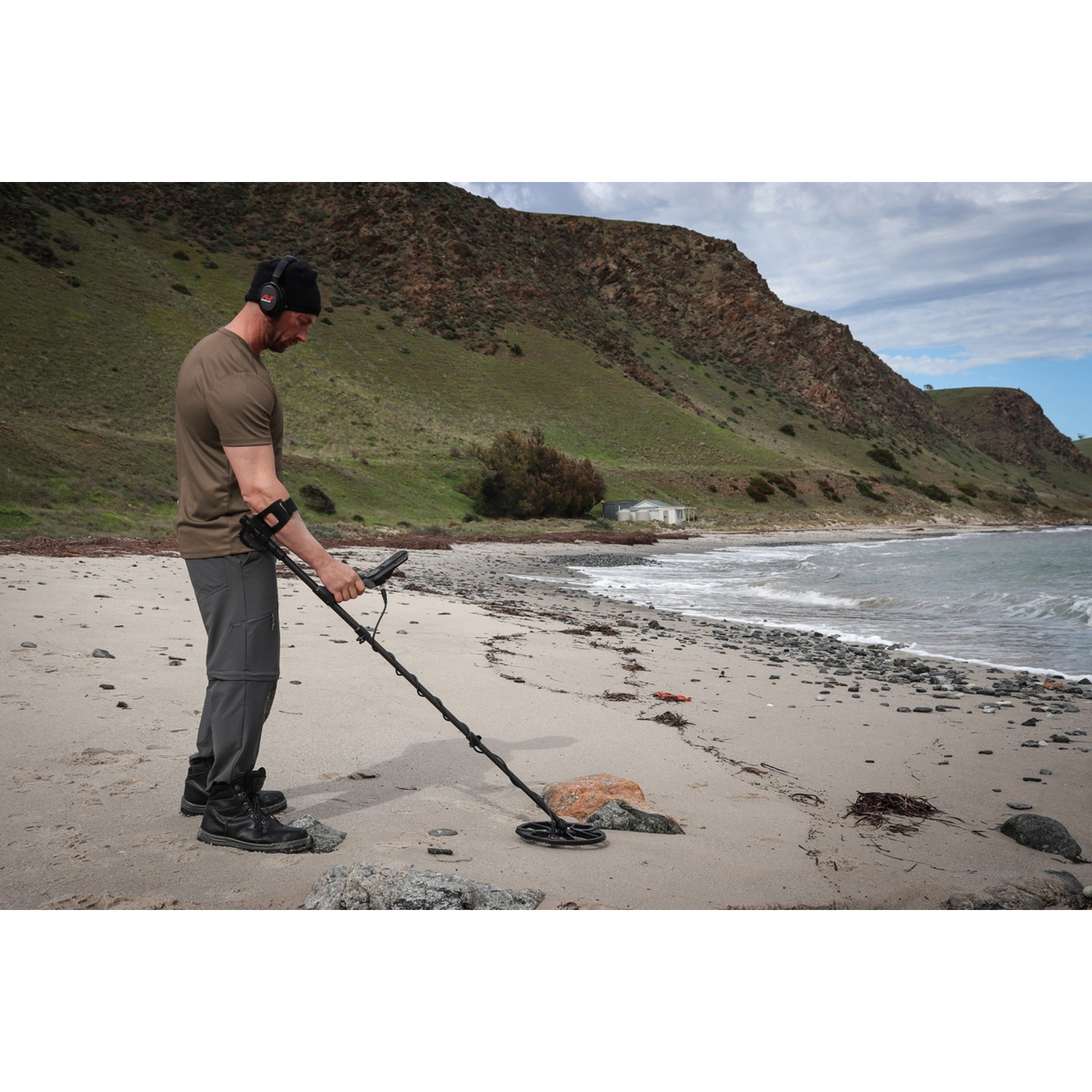 Uomo con cuffie e metal detector su spiaggia sabbiosa, con colline rocciose sullo sfondo.