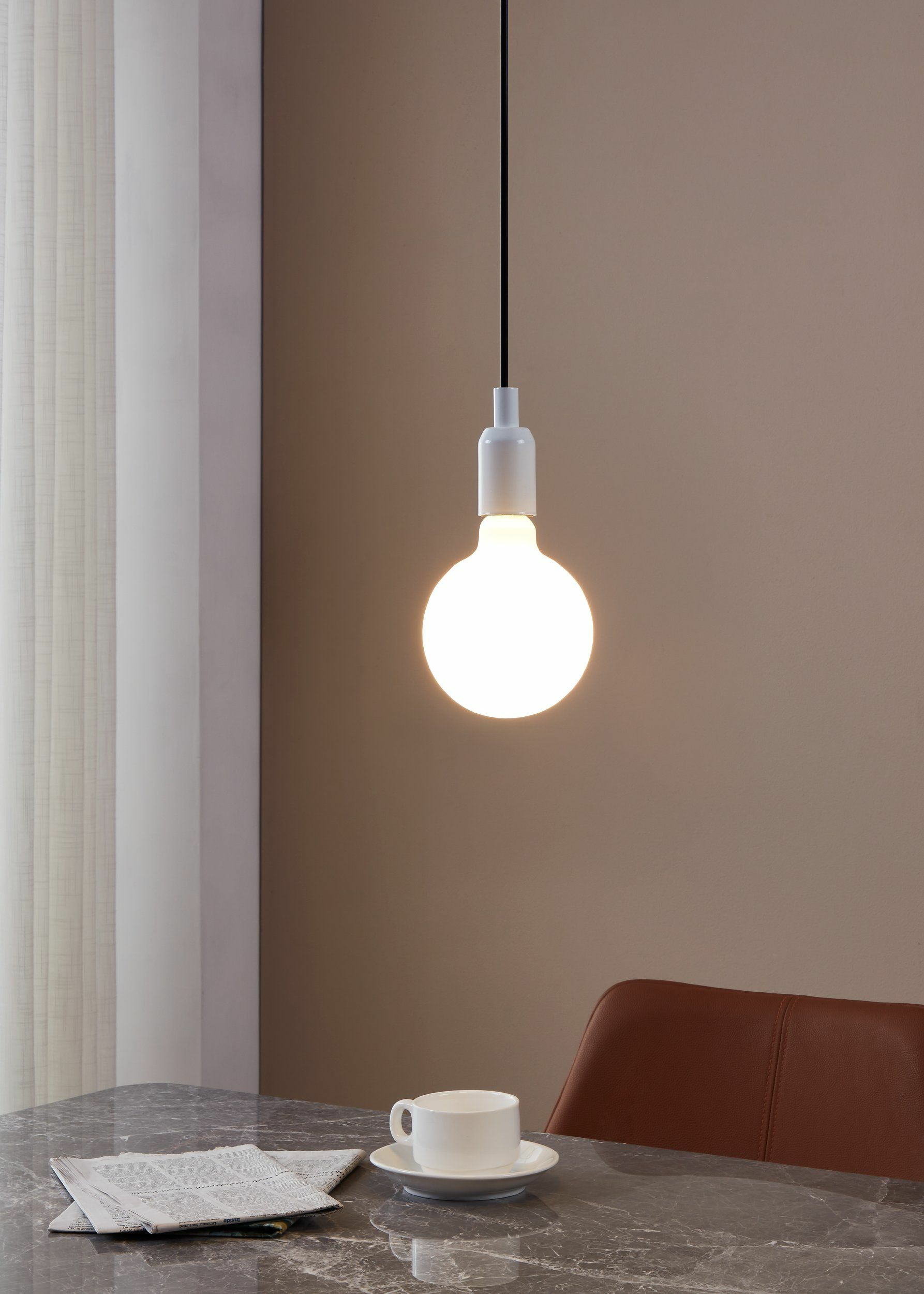 Une lampe suspendue au-dessus d'une table avec journal et tasse. Une chaise marron est visible.