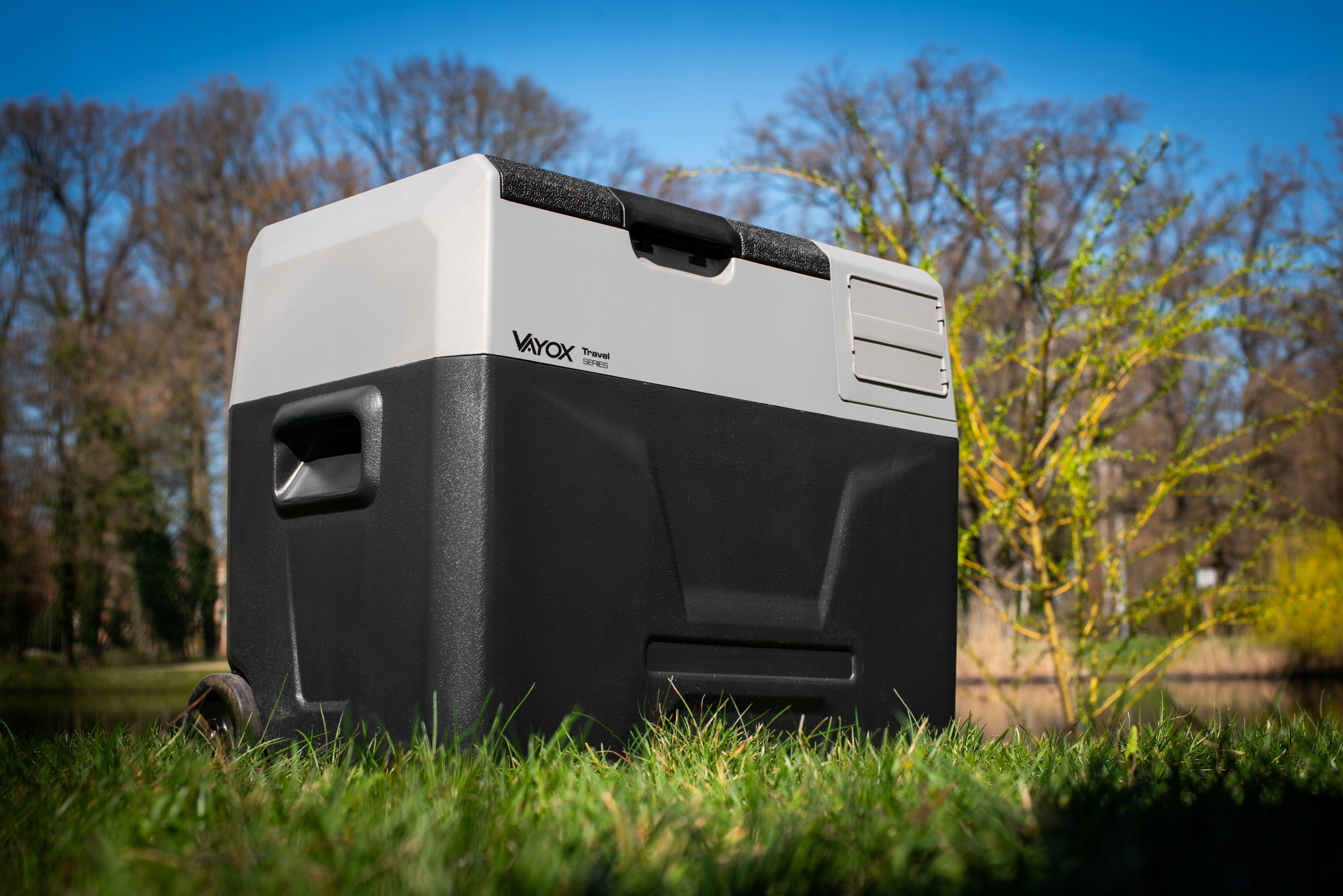Czarno-szara lodówka turystyczna stoi na trawie, z drzewami i niebieskim niebem w tle.