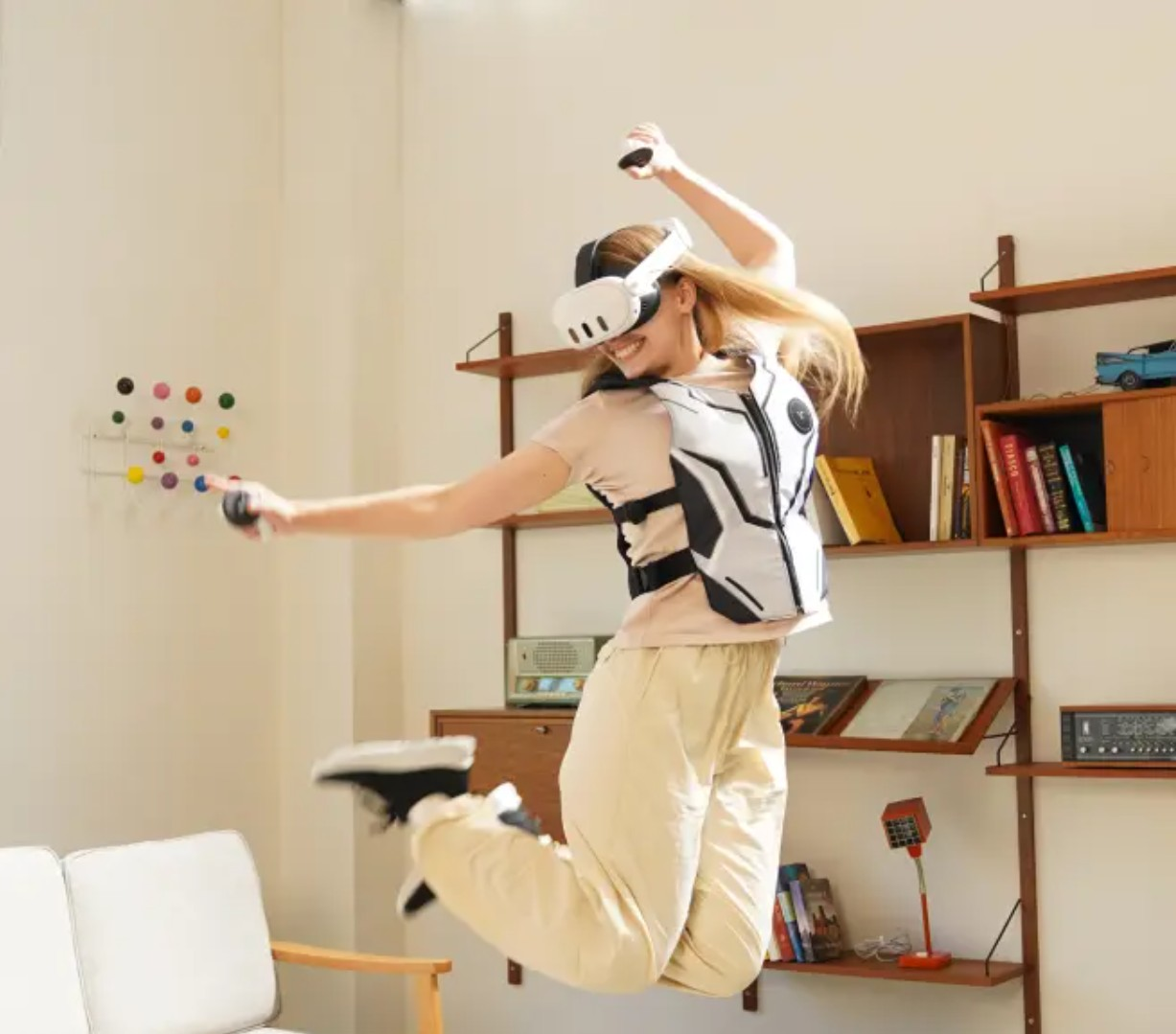 Una persona con casco de realidad virtual y chaleco háptico blanco salta, sosteniendo controladores, en una habitación con estantería y decoración de pared colorida.