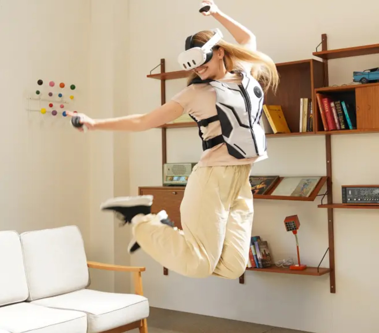 Una mujer con gafas de realidad virtual y chaleco háptico salta en una sala de estar, sosteniendo controladores, con un sofá blanco y estantes detrás.