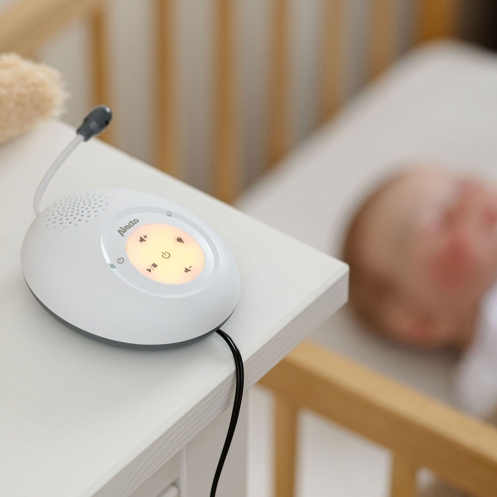Ein weißes Babyfon liegt auf einem Nachttisch neben einem Kinderbett mit einem Baby.