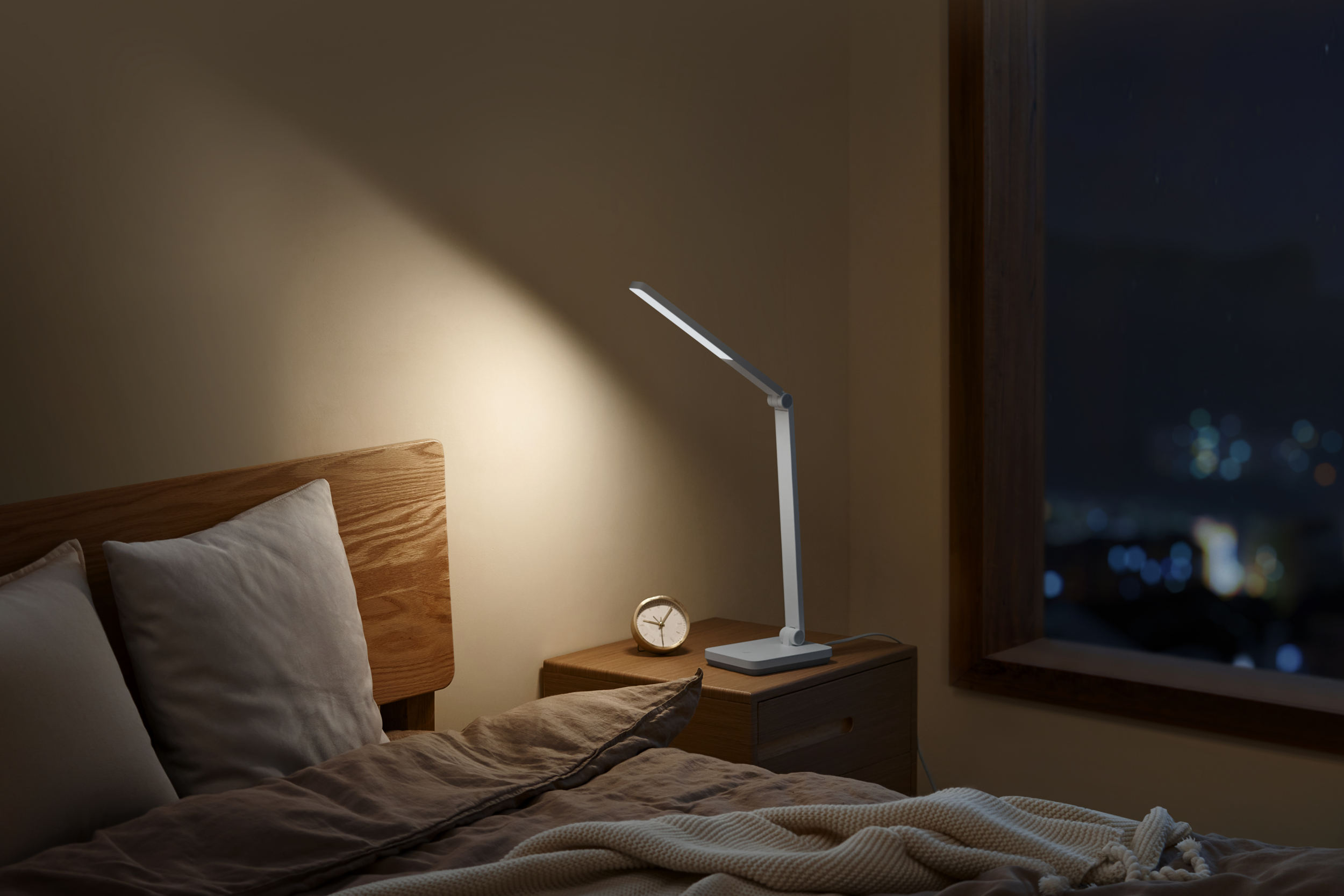 Ein Nachttisch mit Lampe und Wecker neben einem Bett mit Blick auf die nächtliche Stadt.